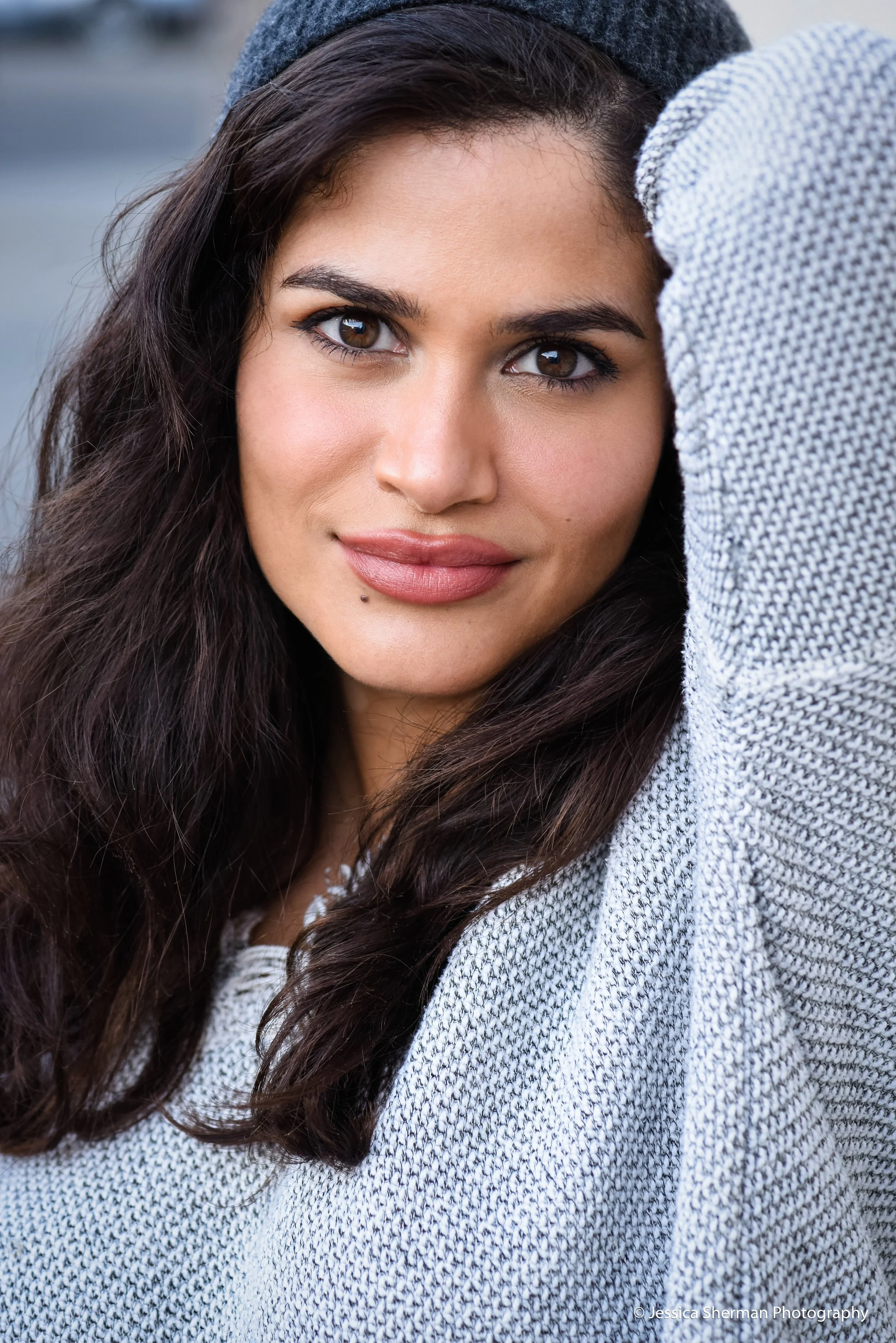 Close-up of a woman with long dark hair, wearing a gray beanie and a light gray sweater, looking at the camera with a slight smile.