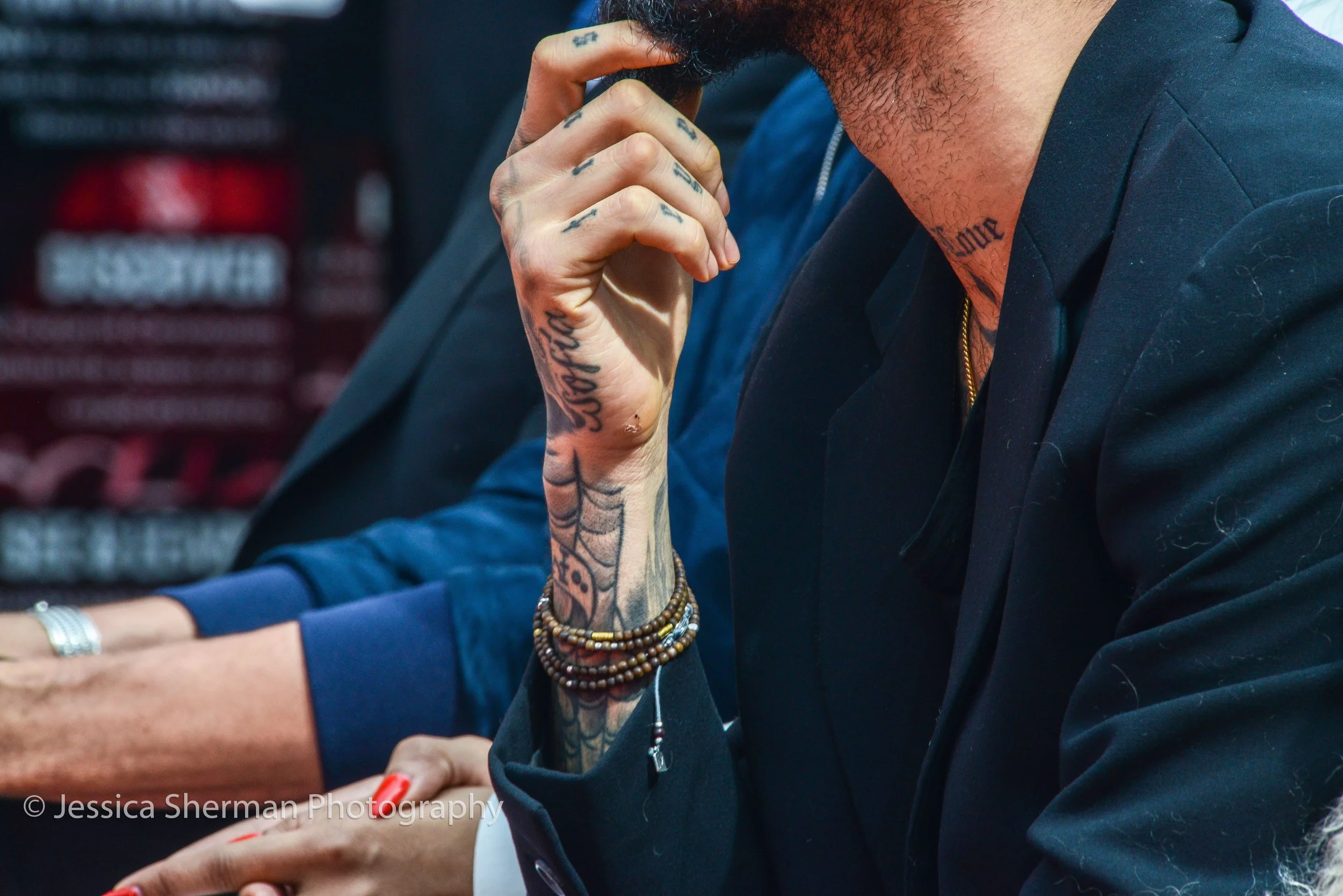 Close-up of a person with tattooed hands and wrist, wearing bracelets and a black shirt, sitting at a table with their hand near their face.