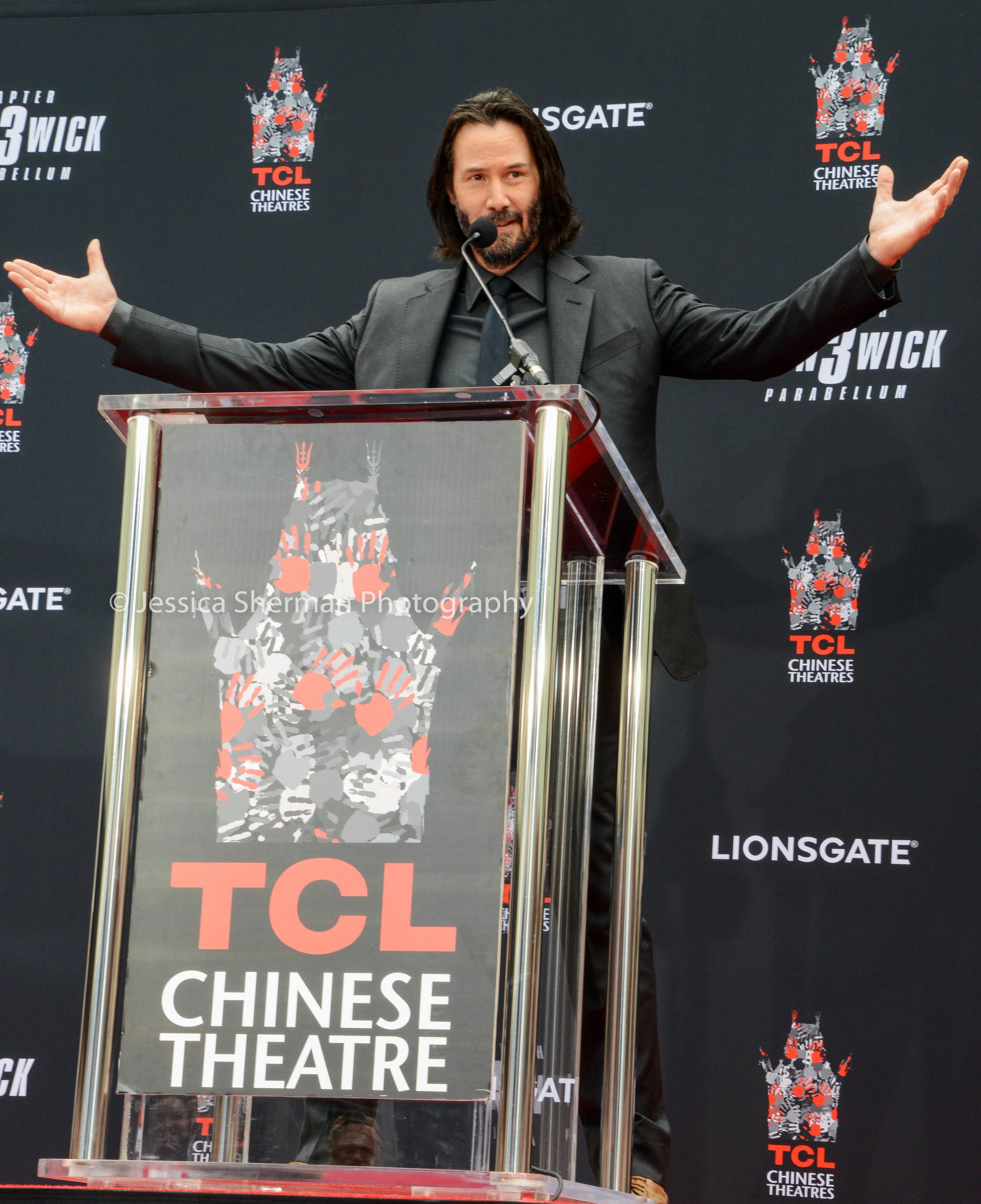 A man with shoulder-length dark hair and a beard dressed in a black suit speaking at a TCL Chinese Theatre event. He is standing behind a transparent podium with the TCL Chinese Theatre logo, with his arms outstretched. The background features TCL Ch