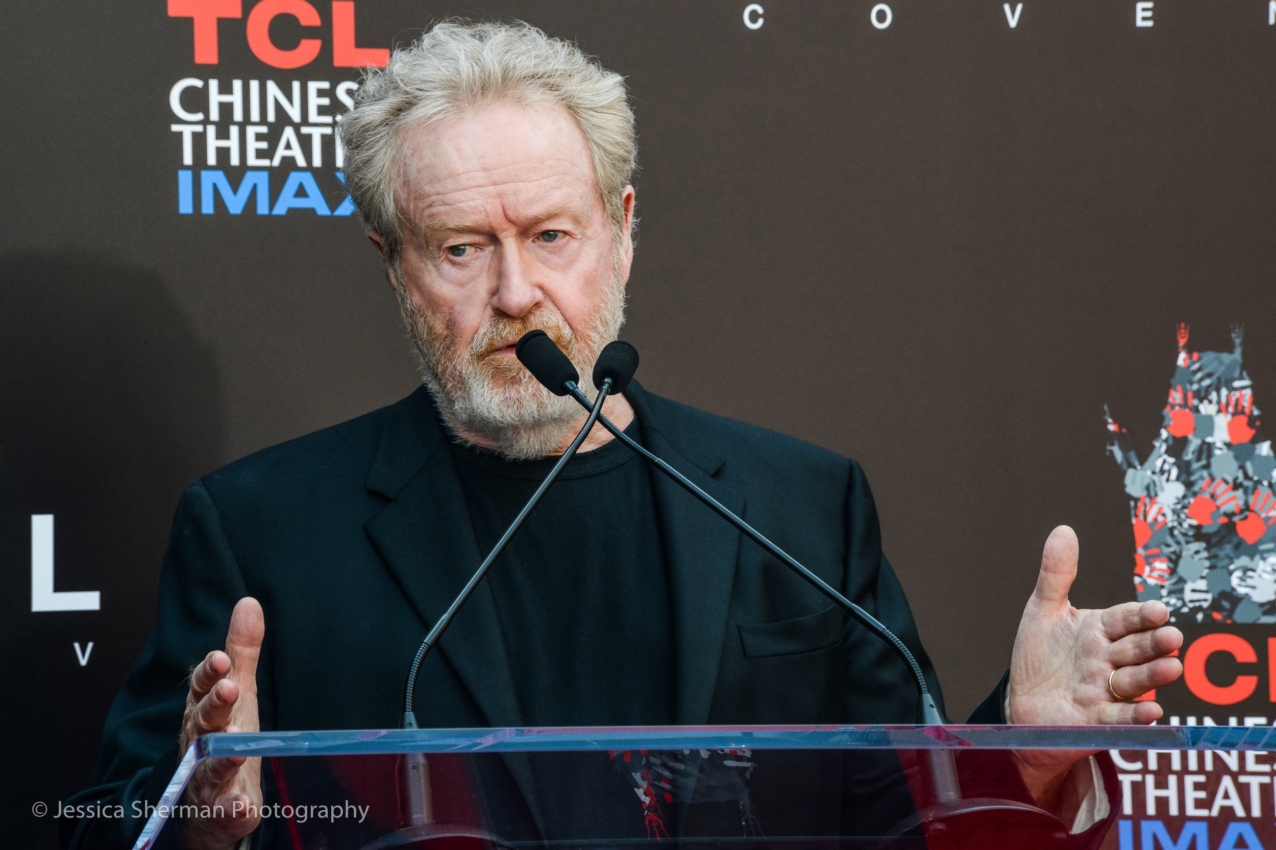 An older man with white hair and a beard speaking at a press conference, standing behind a clear podium, with a dark backdrop displaying the logos of TCL Chinese Theatre and IMAX.