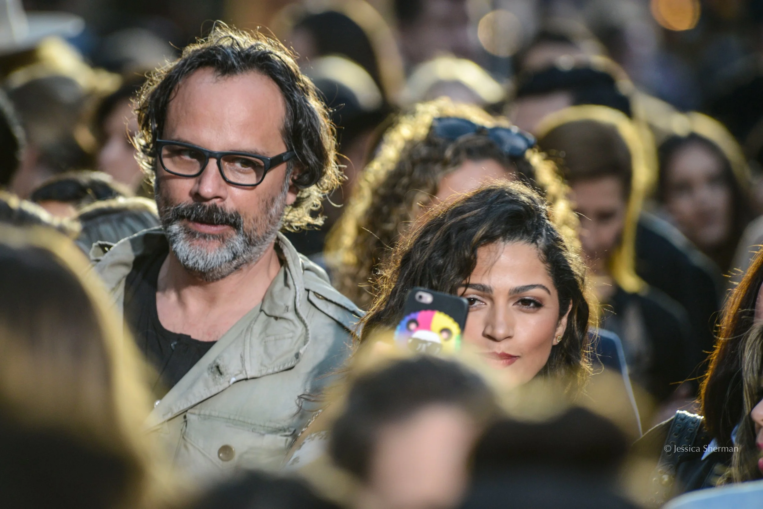 A crowd of people at an outdoor event, with a man with glasses and a beard and a woman with dark hair in the foreground, surrounded by others with some wearing sunglasses and black or yellow clothing.