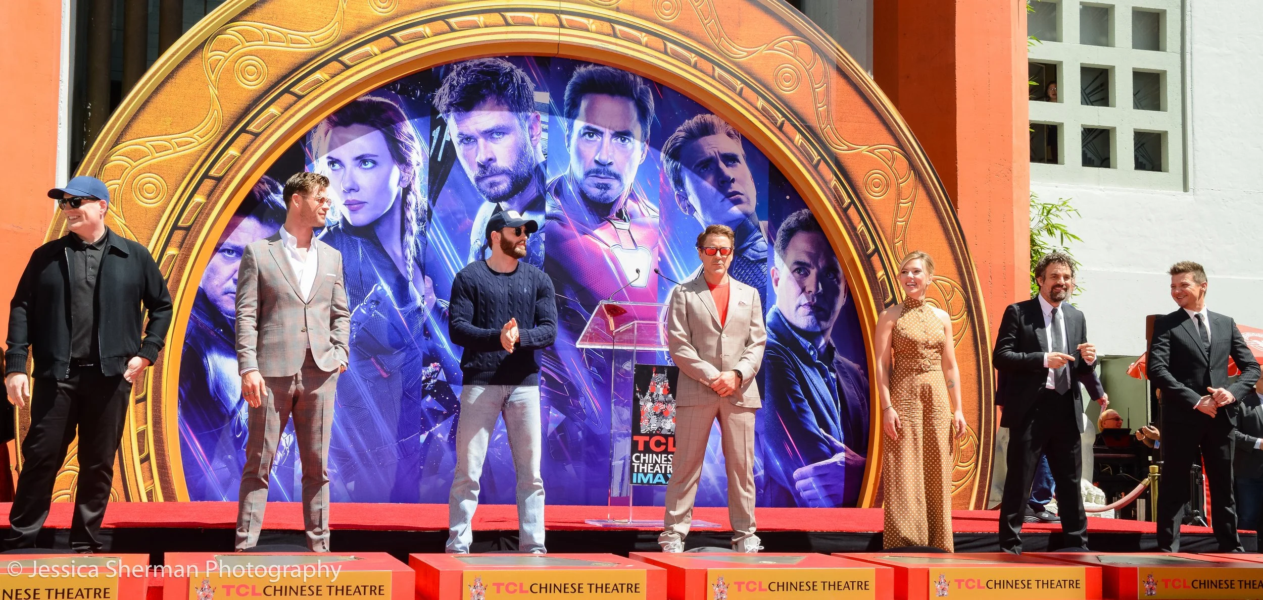 Group of people standing on a stage in front of a large poster of Marvel's Avengers, including Iron Man, Captain America, and Black Widow, at a TCL Chinese Theatre event.