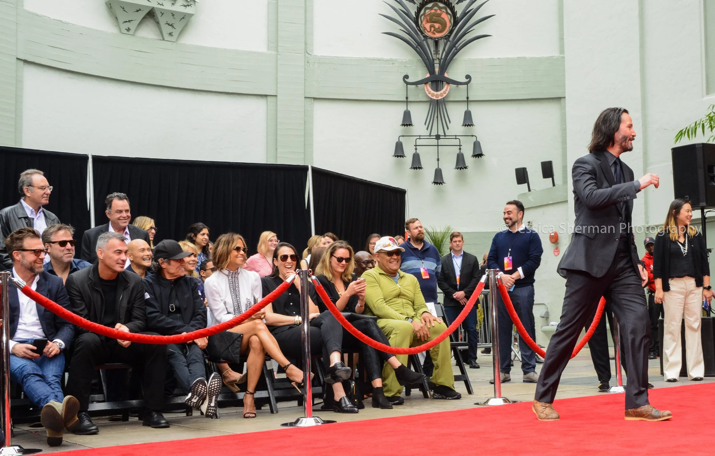A man with long hair and a beard, wearing a dark suit, is walking on a red carpet at an event. Behind him, several smiling audience members sit and stand, some taking photos, in a large indoor space with a green painted wall and large black decor han