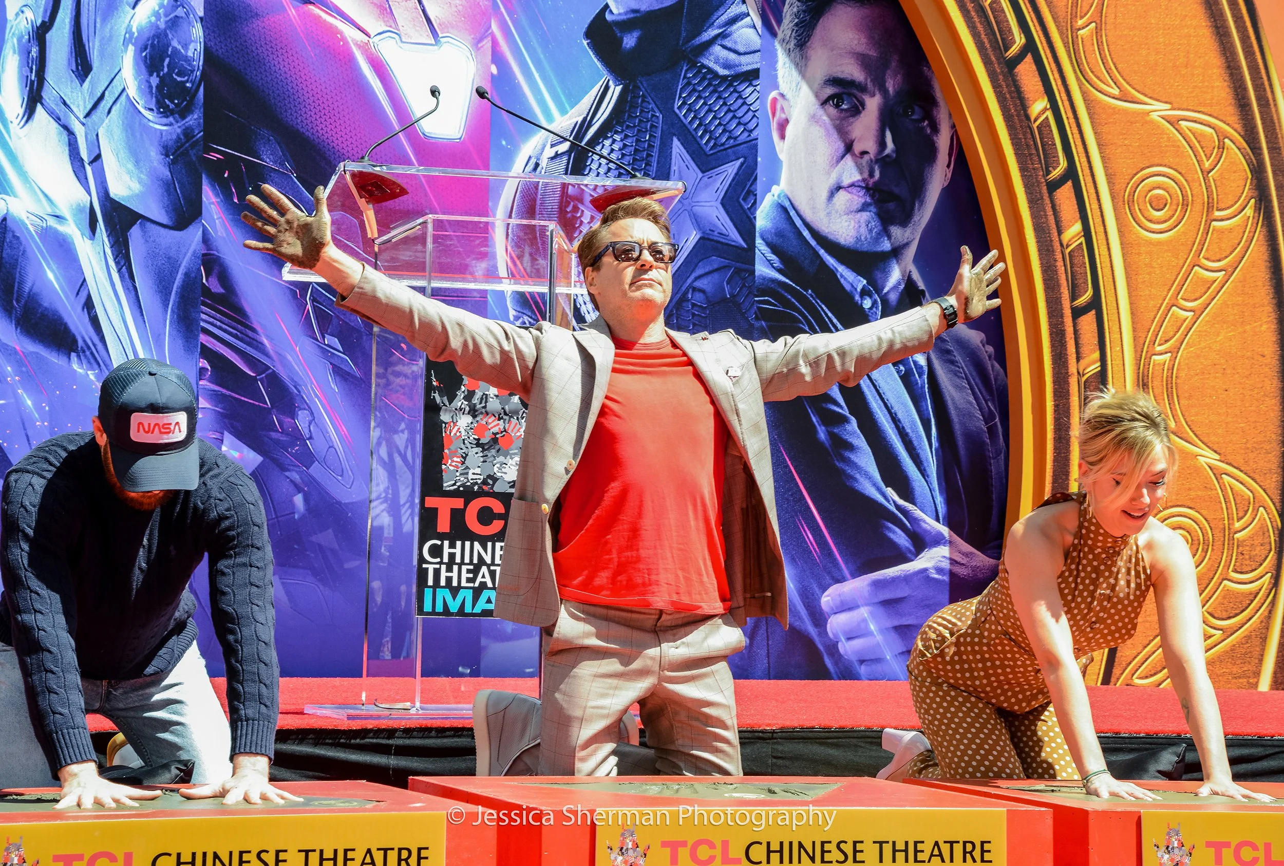 Three standees participating in an event on stage at the TCL Chinese Theatre, with a large movie poster in the background depicting characters from a superhero film. The person in the center is kneeling with arms outstretched, wearing sunglasses and 