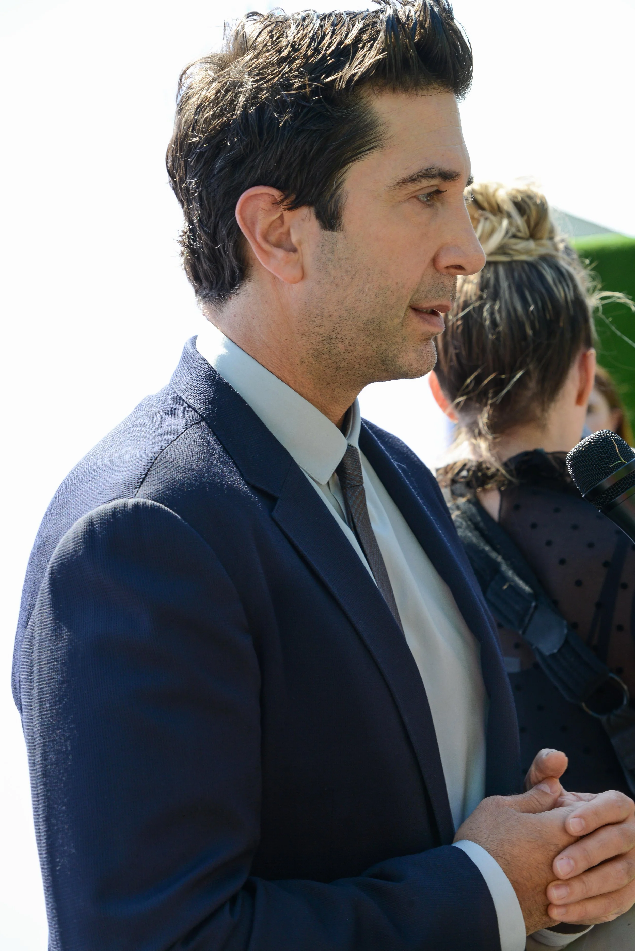 A man in a dark suit and tie is speaking outdoors, with a woman holding a microphone in the background.