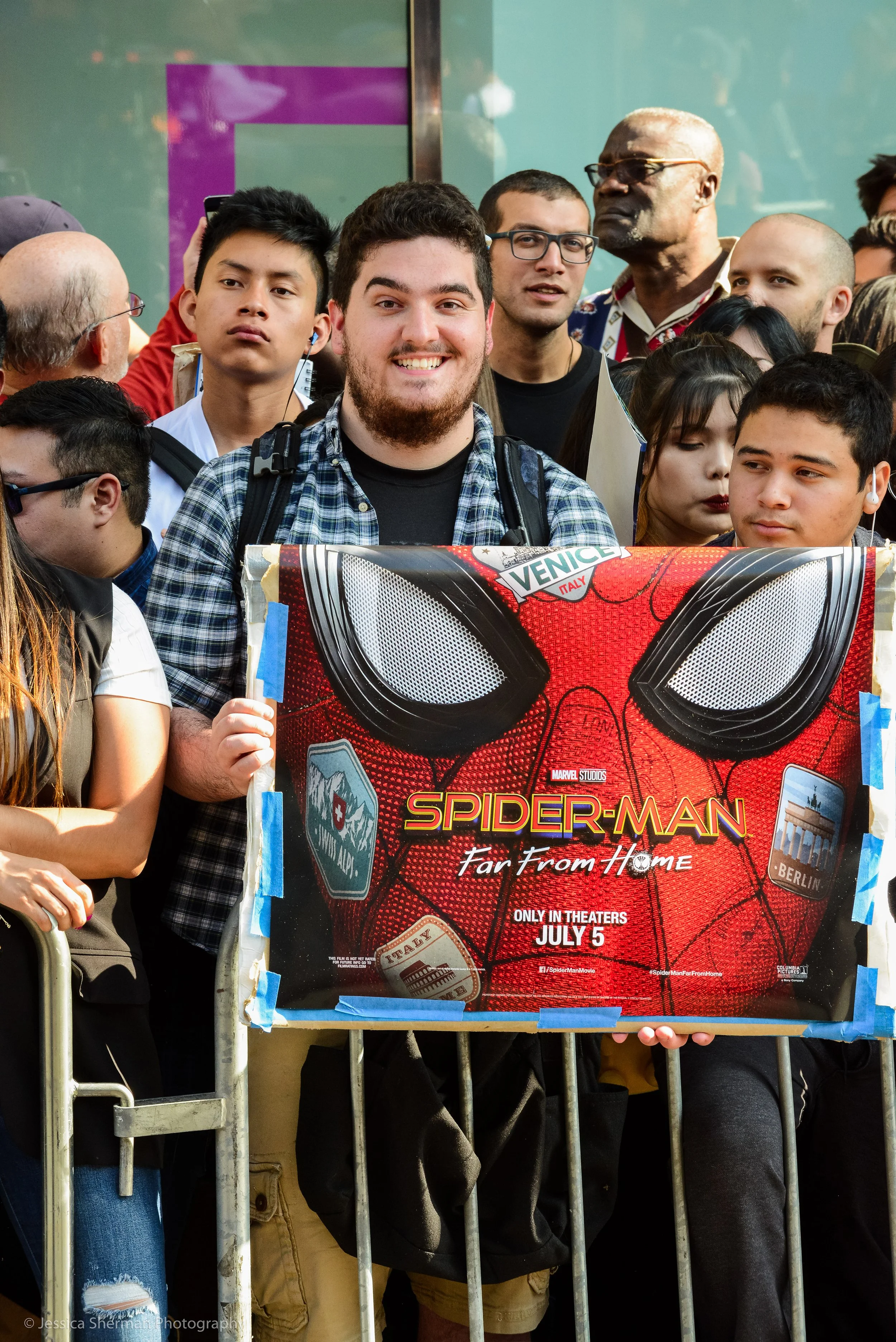People standing behind a barricade at a Spider-Man movie premiere, holding a promotional poster for 'Spider-Man: Far From Home' with a red and black design, featuring Spider-Man's mask and release date of July 5.