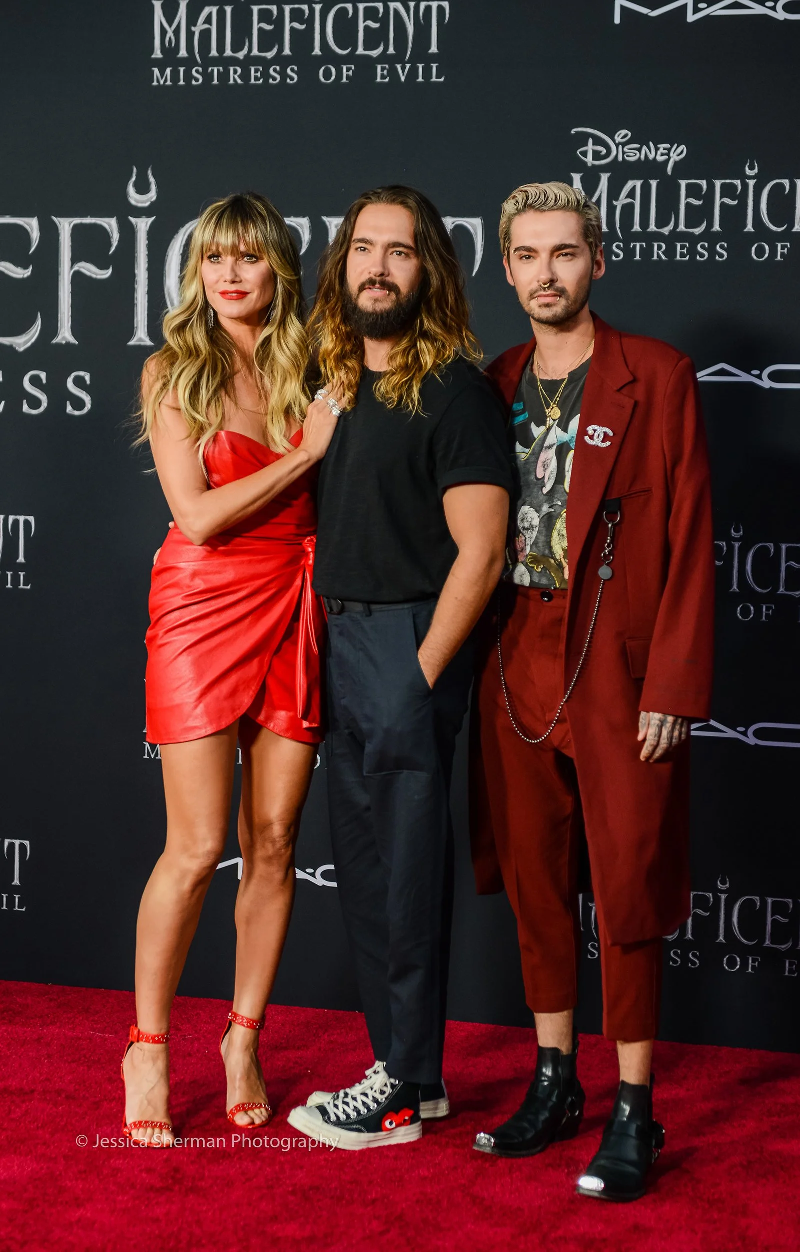 Three people posing on a red carpet in front of a black backdrop with "Maleficent Mistress of Evil" and "Disney" logos. One woman in a red dress, a man in black with long hair, and another man in a red suit with tattoos.