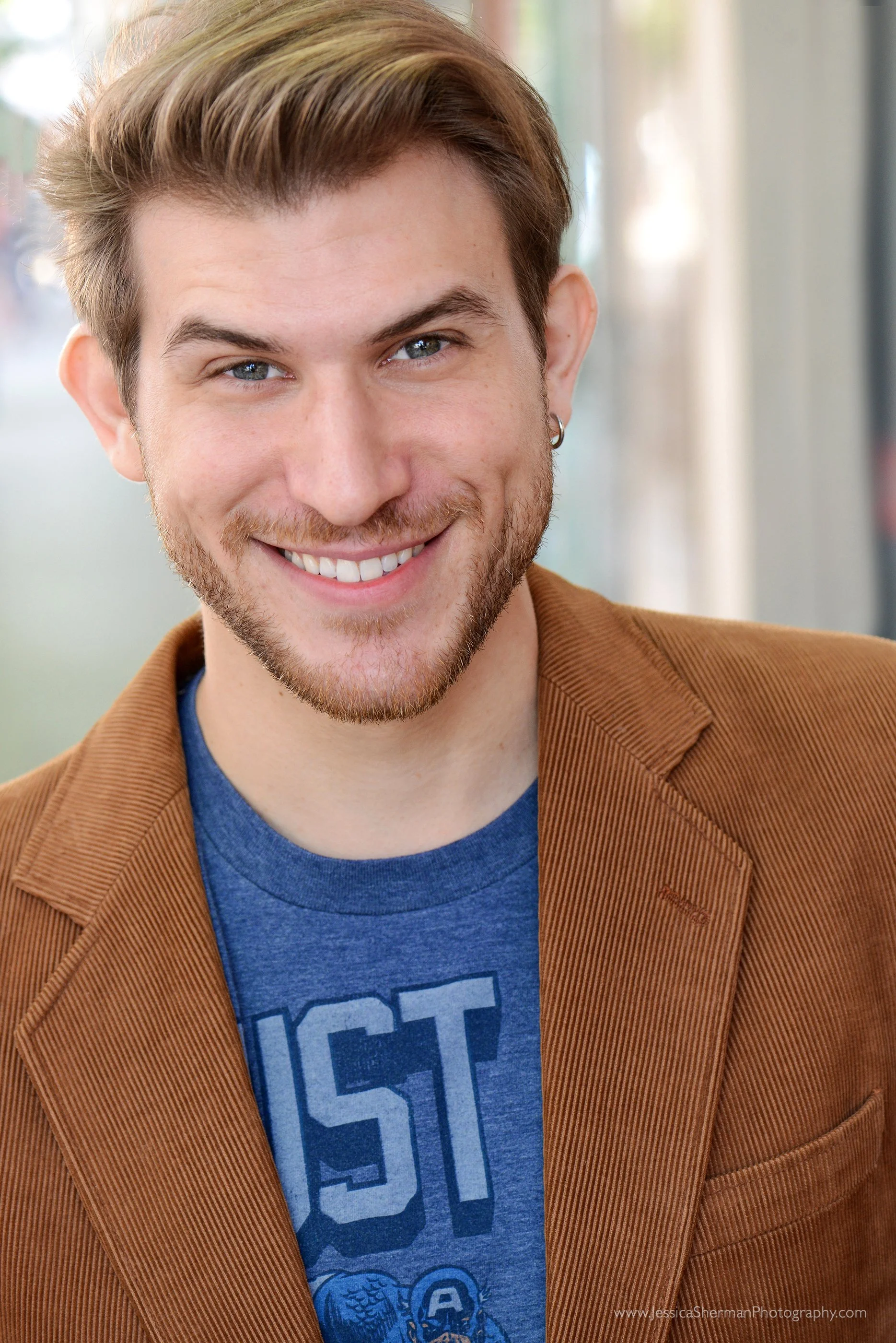 Close-up portrait of a young man with styled brown hair, blue eyes, and a smile. He's wearing a brown jacket over a blue T-shirt with a graphic design and is in an indoor setting with blurred background.