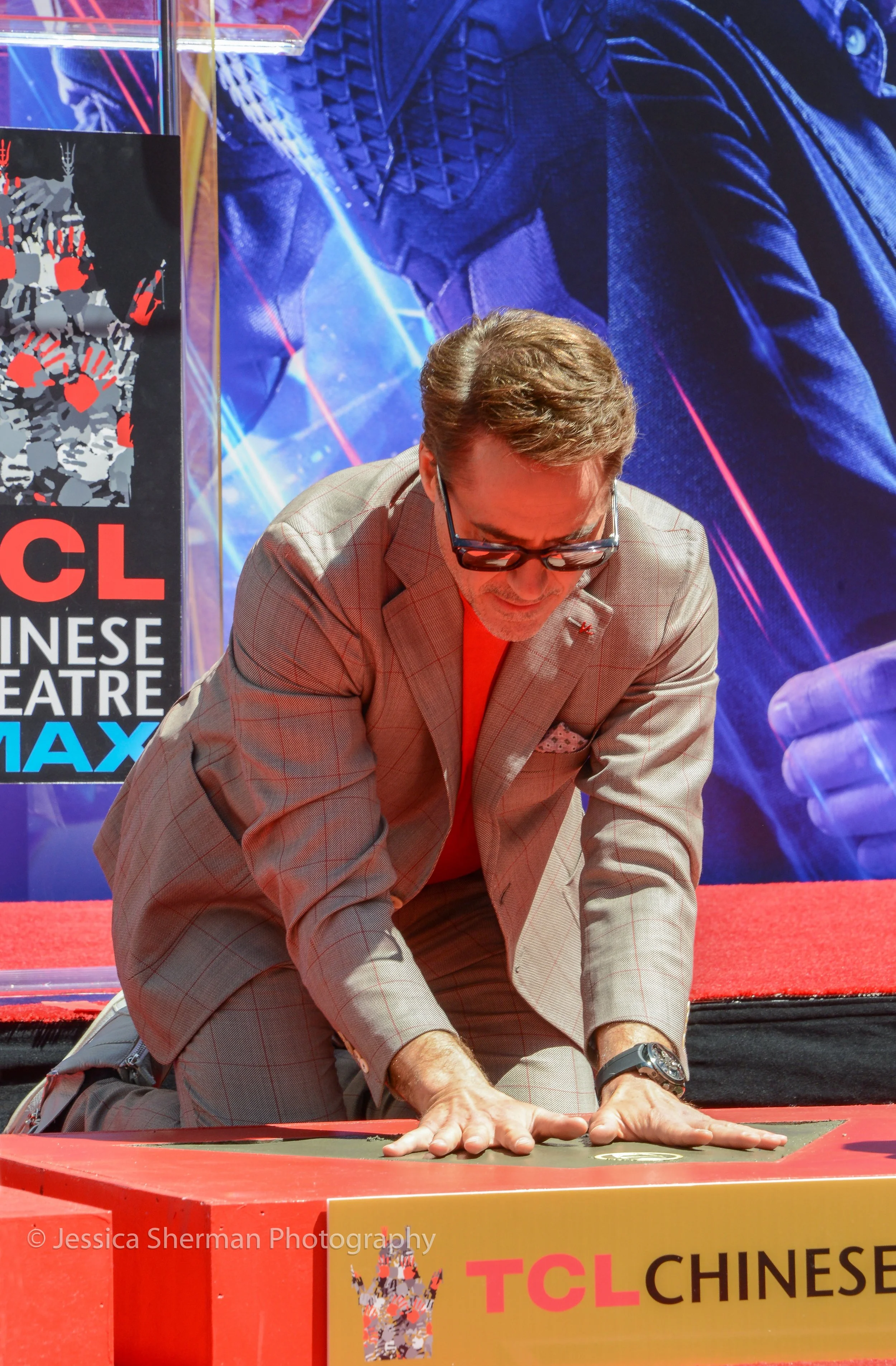 A man in a beige plaid suit and glasses kneeling on the red carpet with both hands on a raised platform, participating in a game of mahjong, at the TCL Chinese Theatre during a MAX event.