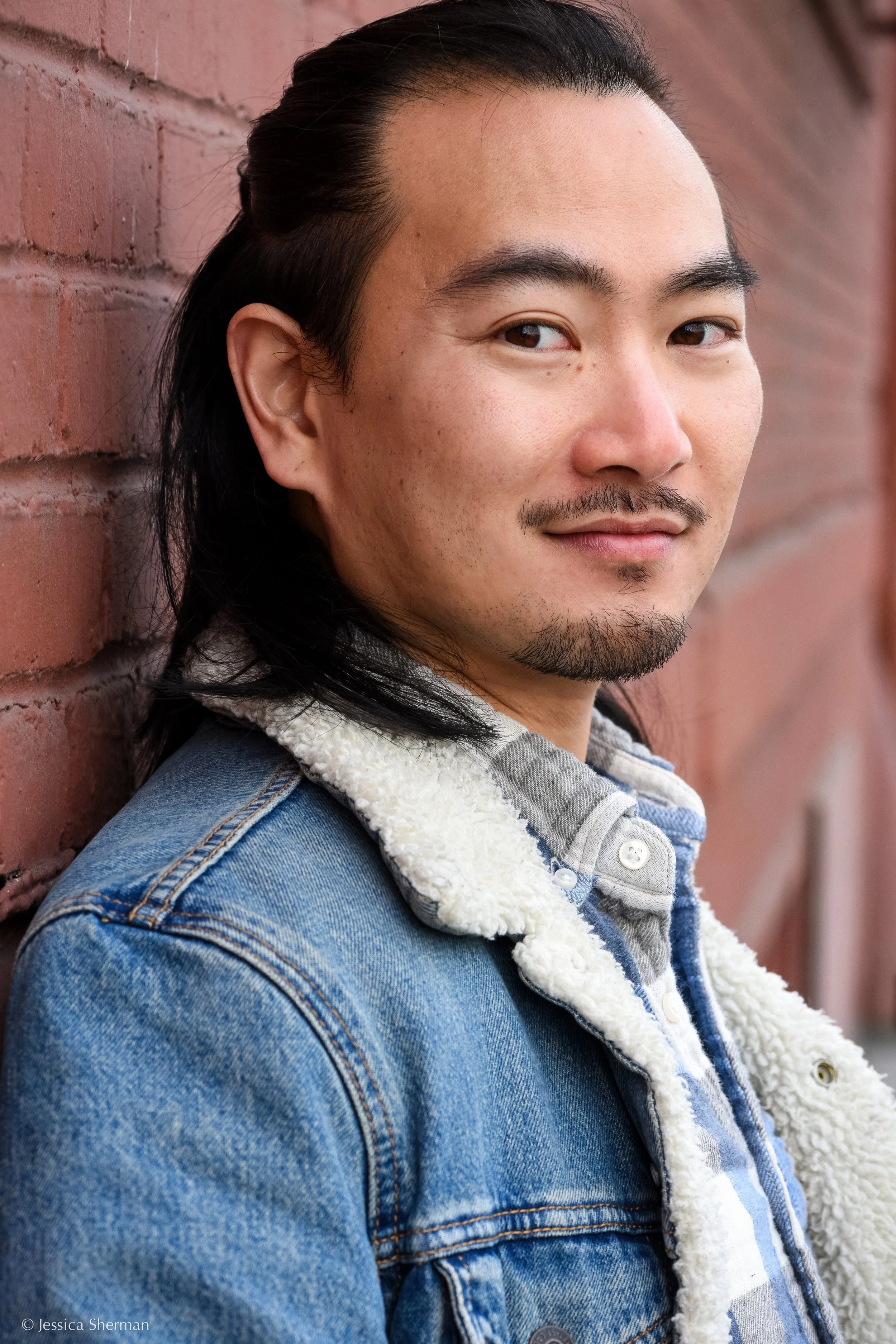 A man with long black hair, a goatee, and mustache, wearing a denim jacket with a sherpa lining, leaning against a red brick wall, looking at the camera with a slight smile.