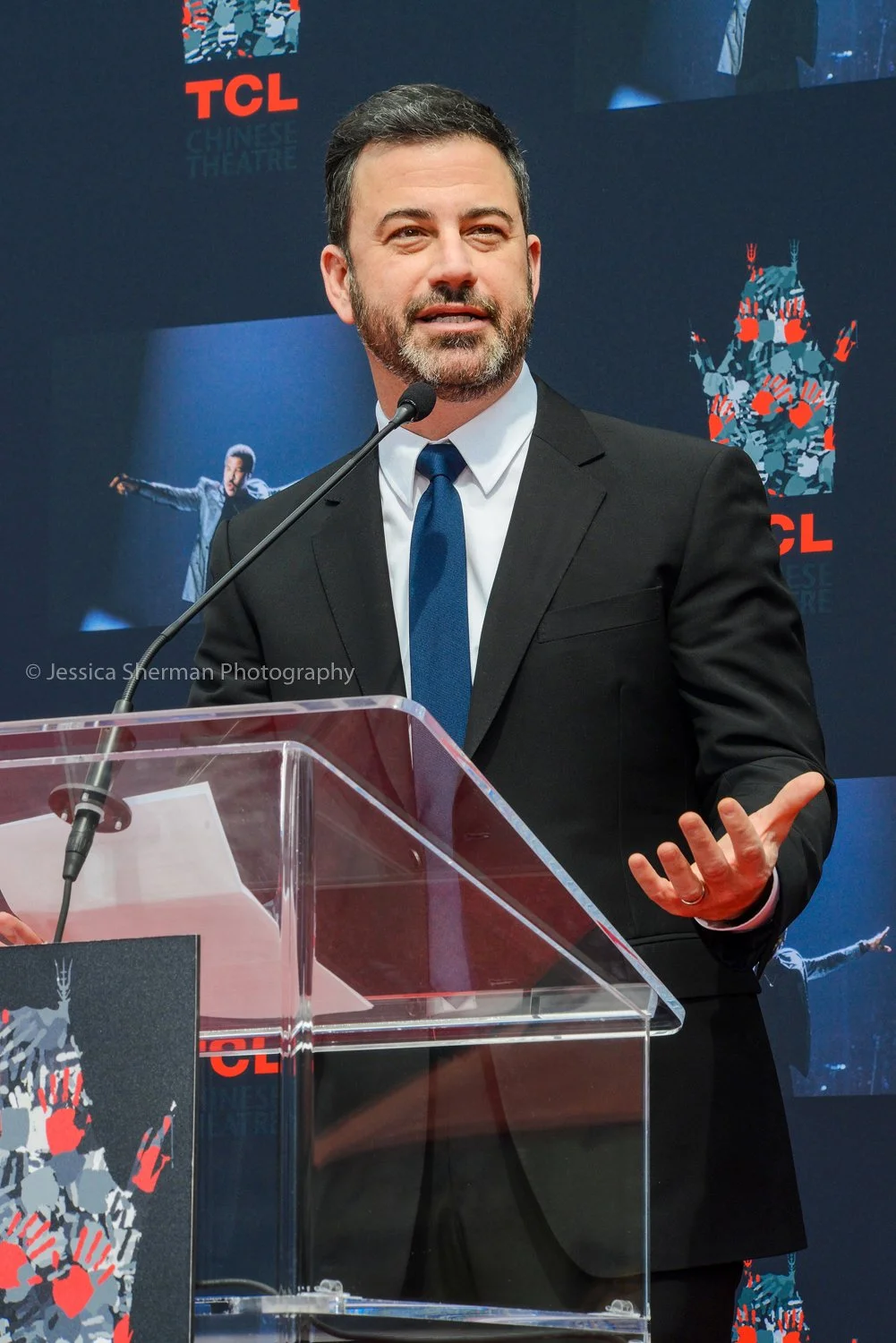 A man wearing a black suit, white shirt, and blue tie speaking at a podium during an event, with a backdrop showing the TCL Chinese Theatre logo and images from a performance on a large screen behind him.