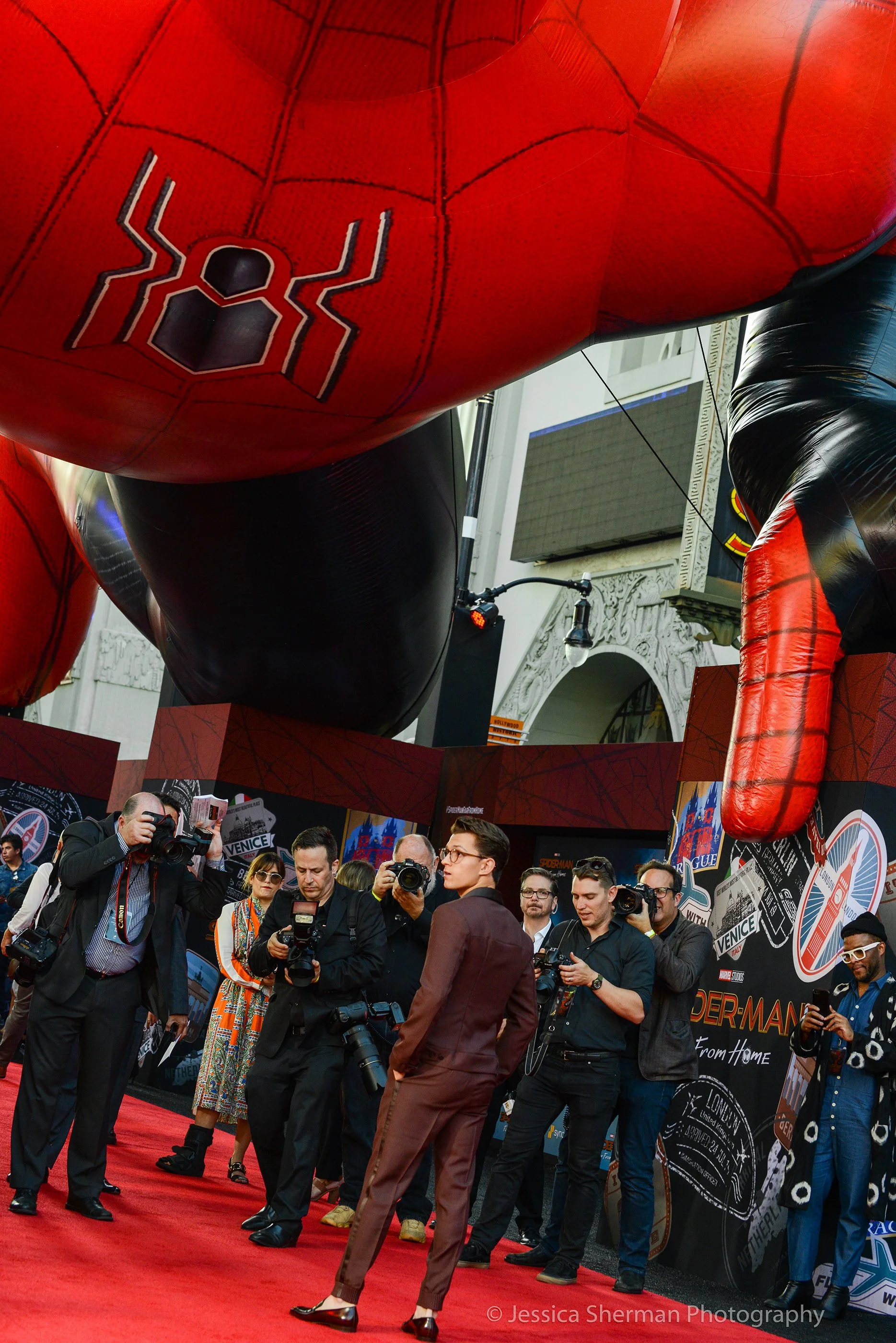 A group of photographers and media personnel take photos and videos at a Spider-Man themed event, with a large Spider-Man inflatable figure hanging overhead.