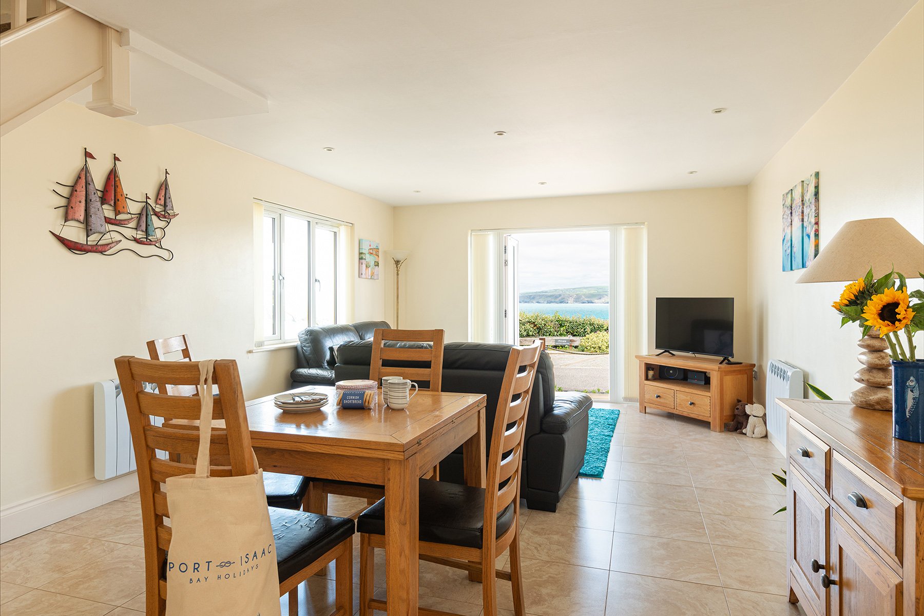 Living room with a dining table, black sofa, TV on a wooden stand, and large windows with a view of the sea and the coastline of port isaac bay