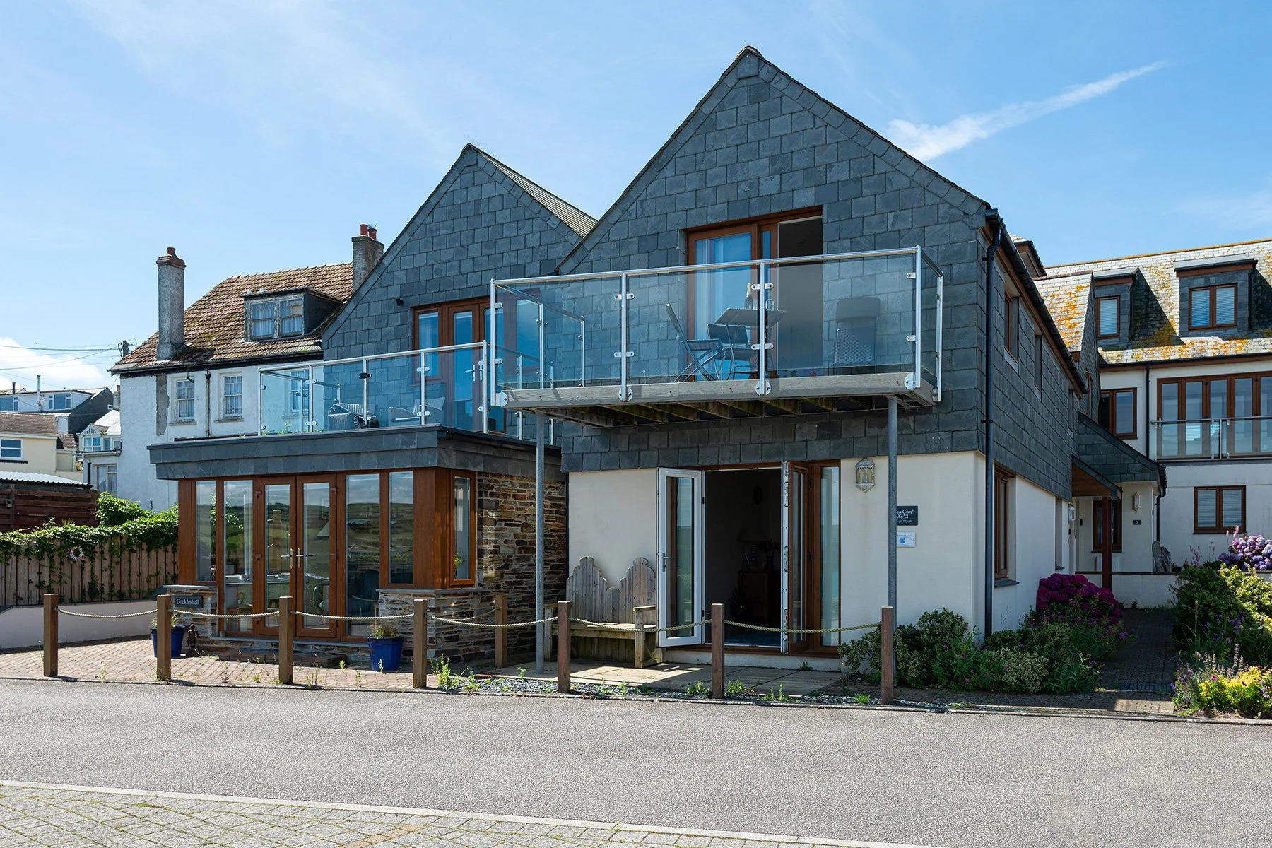 A modern house with gray slate siding and large glass doors, featuring a glass balcony with seating on the upper floor. The house has wooden window frames and a small garden with flowers in front. The street in front is paved and lined with a wooden fence.