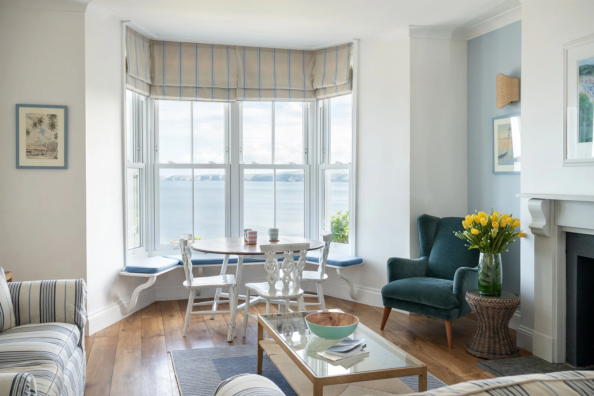 Living room with large bay window overlooking water, with a small round dining table and four chairs, a blue armchair with a bouquet of yellow flowers, a striped sofa, and artwork on the walls.