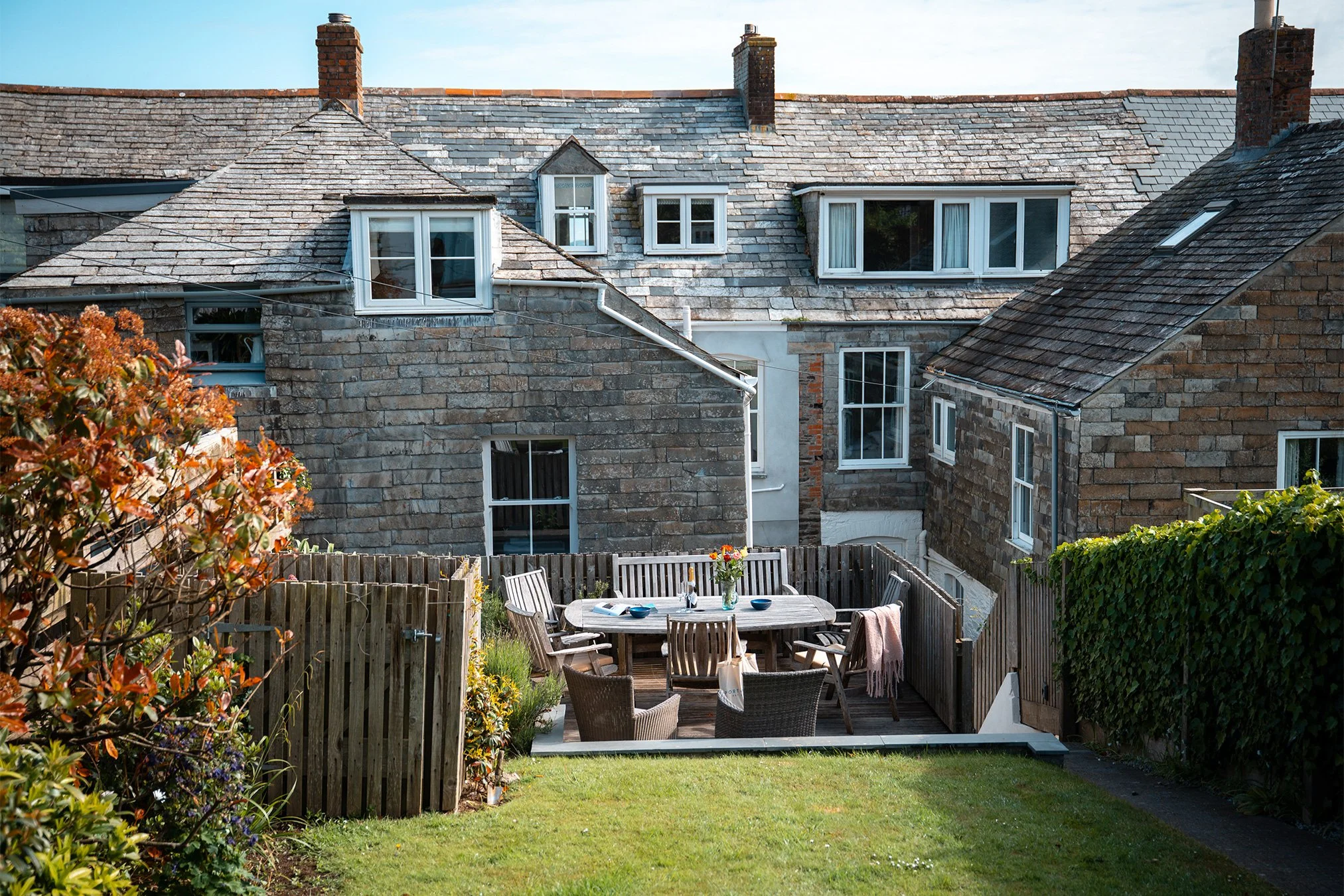Backyard outdoor patio with wooden dining table and chairs, surrounded by a wooden fence, in front of a stone house with multiple windows and a slate roof. There are plants and shrubs along the fence and a grassy lawn.
