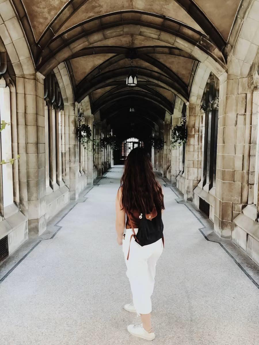 A young female student wearing a black backpack walking through a Gothic-style college hallway built of stone.