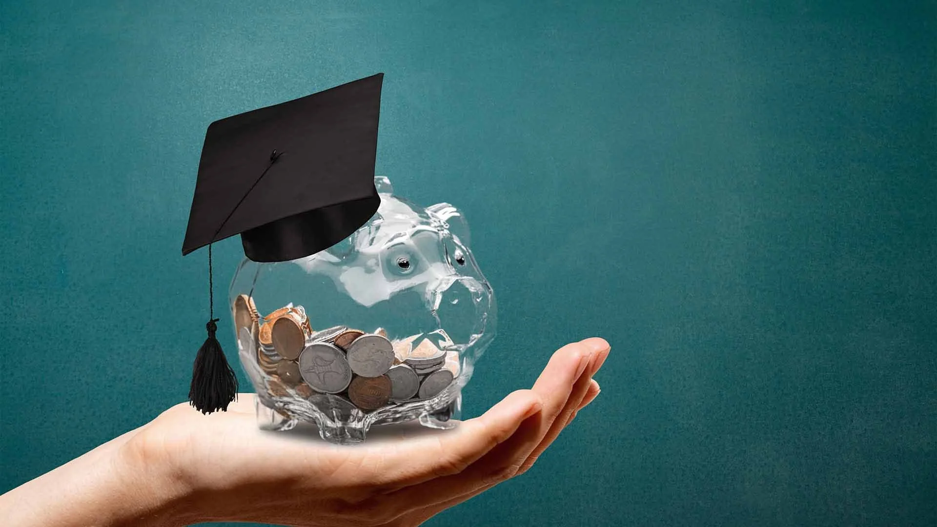 A hand holding a glass piggy bank, filled with coins, decorated with googly eyes, and wearing a black graduation cap, against a green background.