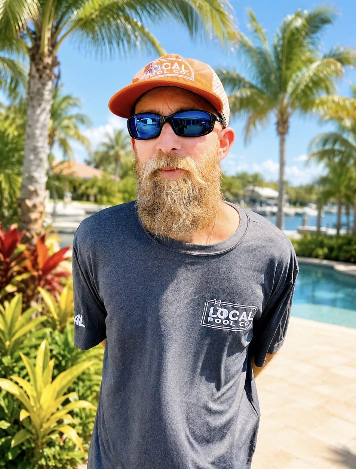 A man with a beard wearing sunglasses, an orange cap with a logo, and a gray T-shirt with a logo, standing in front of a blue corrugated metal wall.