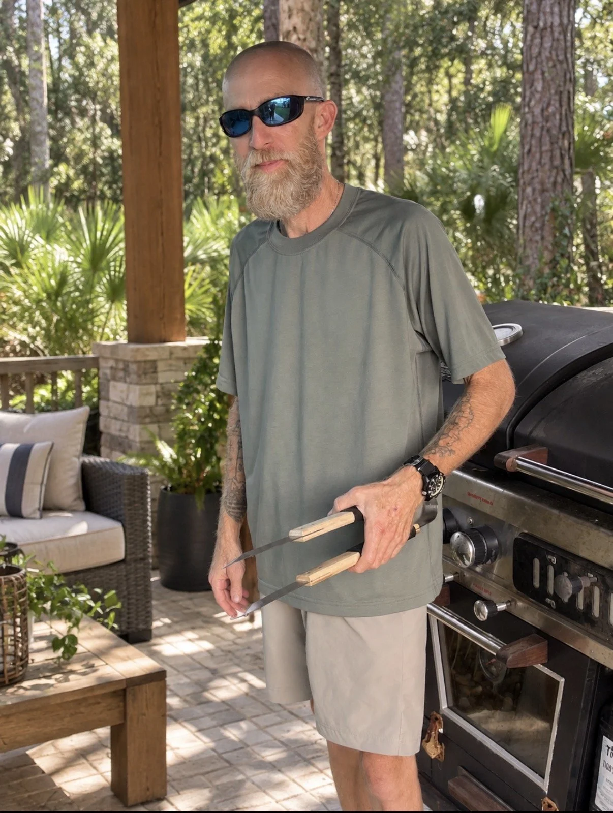 A man with a beard wearing sunglasses, an orange cap with a logo, and a gray T-shirt with a logo, standing in front of a blue corrugated metal wall.