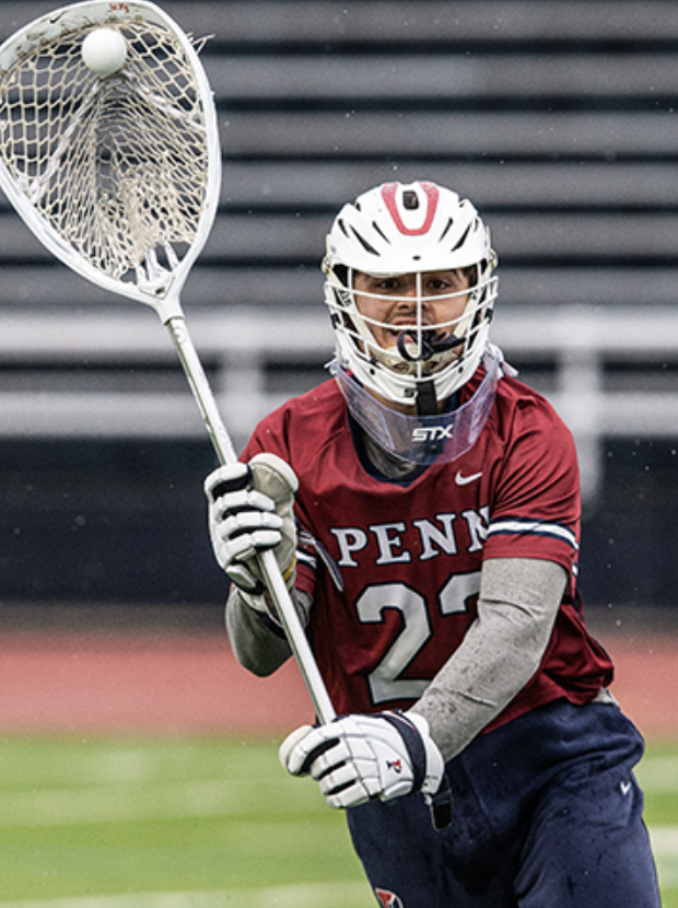A male lacrosse player wearing a Penn State uniform, helmet, and gloves, preparing to catch a ball with his lacrosse stick on a field.