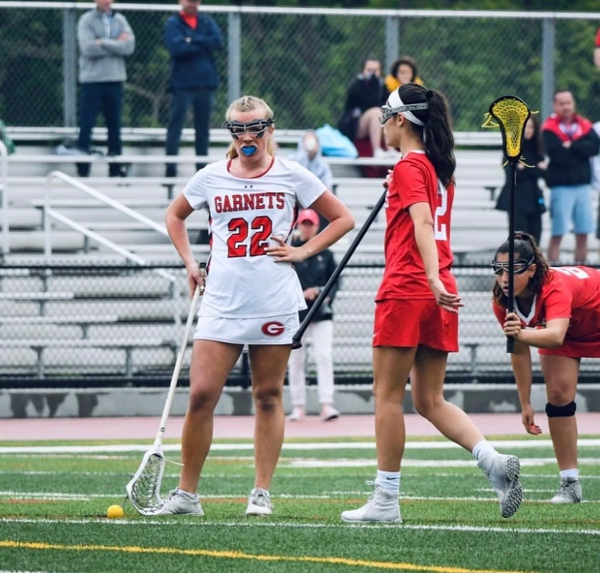 Two female lacrosse players wearing uniforms and goggles on a field, with one speaking and the other standing with a lacrosse stick. Spectators sit in the background on bleachers.