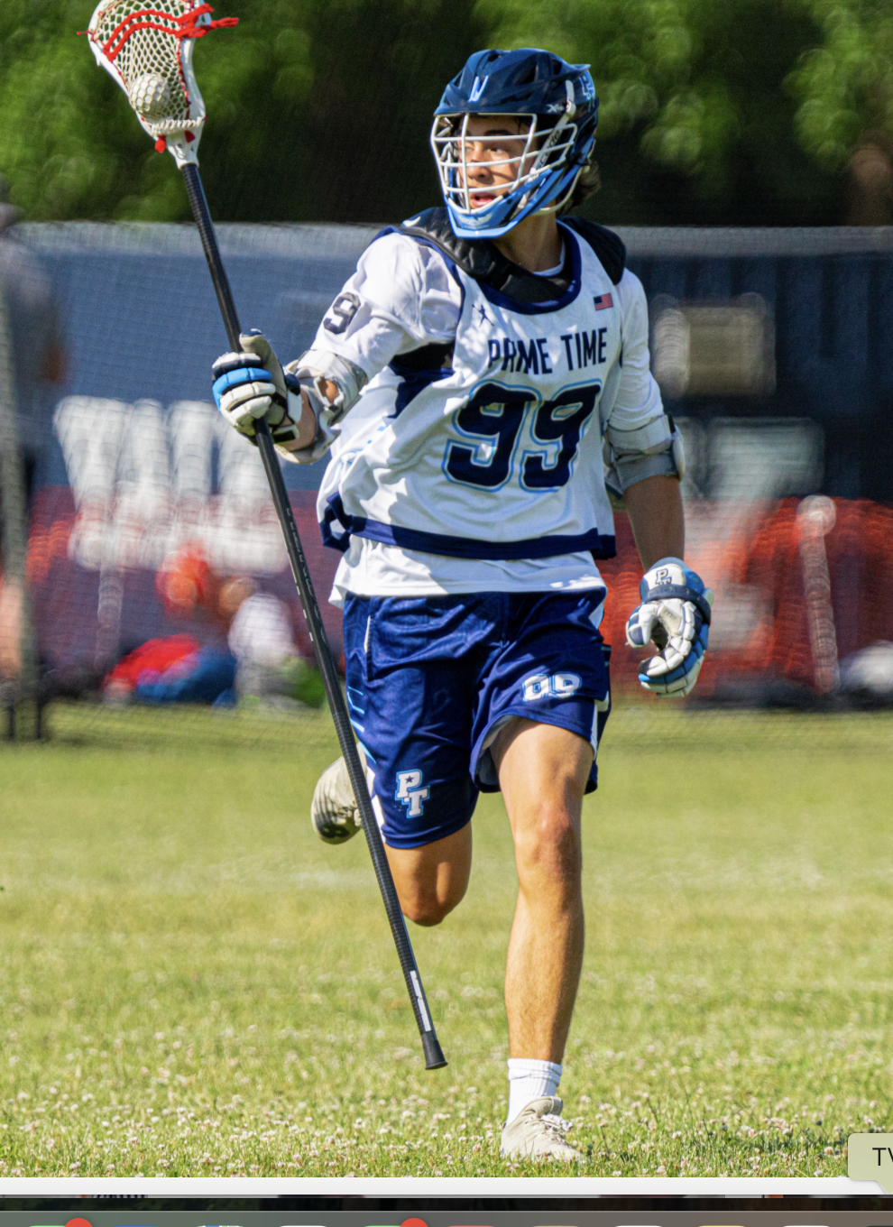 Lacrosse player wearing a blue helmet and white jersey with number 93, running on a grassy field with a lacrosse stick, during daytime.