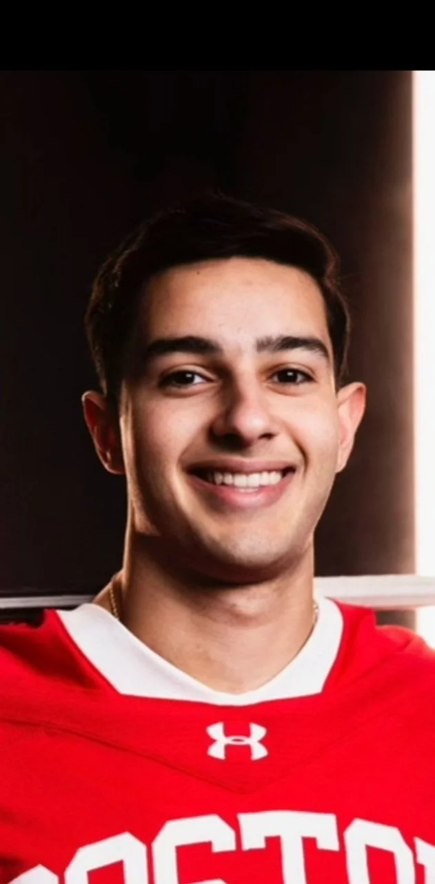 A young man with short dark hair, smiling, wearing a red sports jersey with white trim and white letters, standing indoors with a black background and a vertical light strip on the right side.