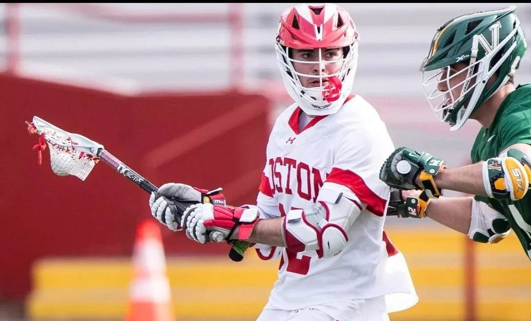 Lacrosse players in game action, one in a white and red uniform holding a lacrosse stick, and the other in a green uniform grabbing the player's arm.