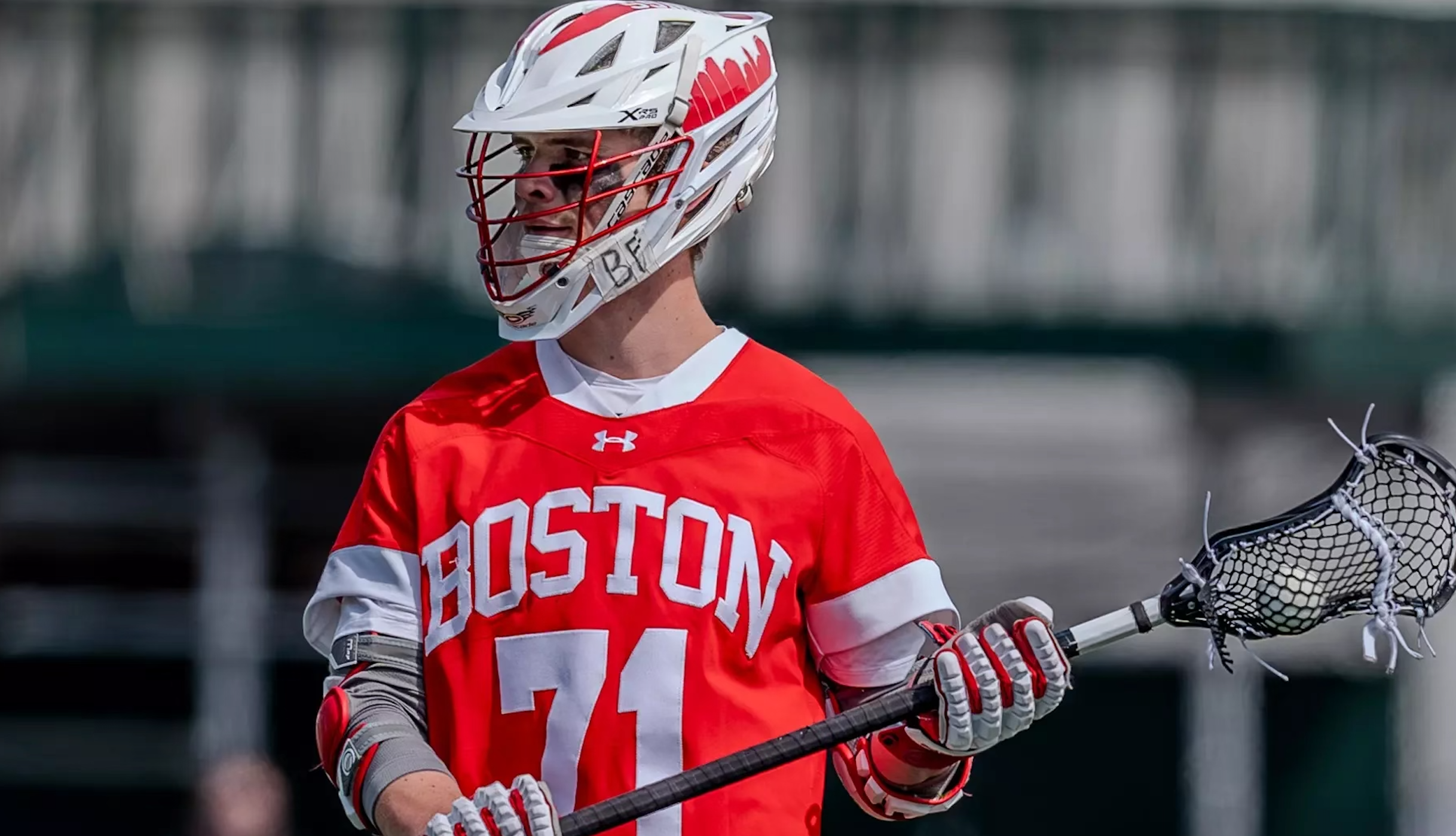 A lacrosse player wearing a red Boston jersey with the number 71, a white helmet, and gray gloves, holding a lacrosse stick on the field.