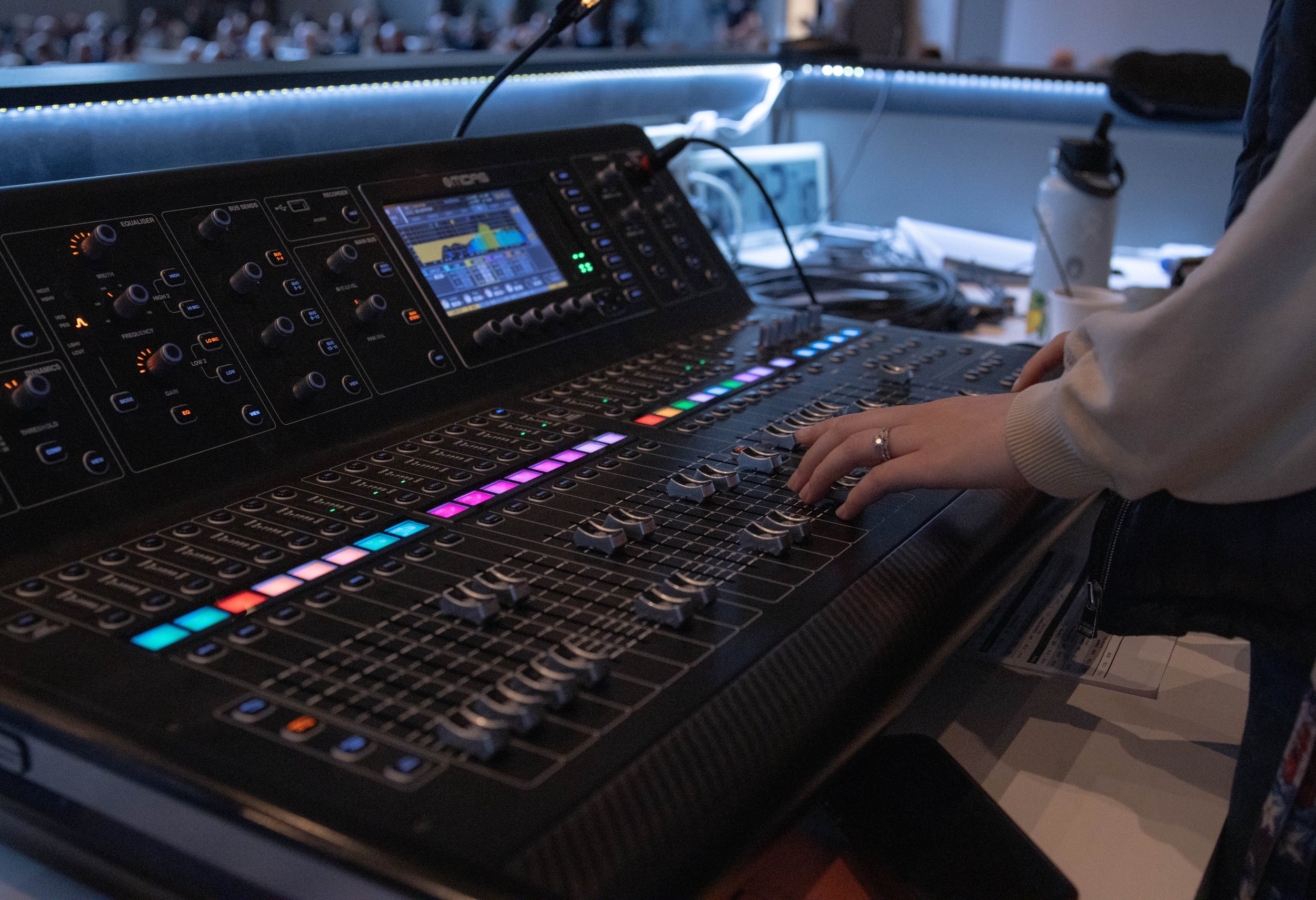 Person adjusting audio mixing console with multiple sliders and illuminated controls in a studio setting.