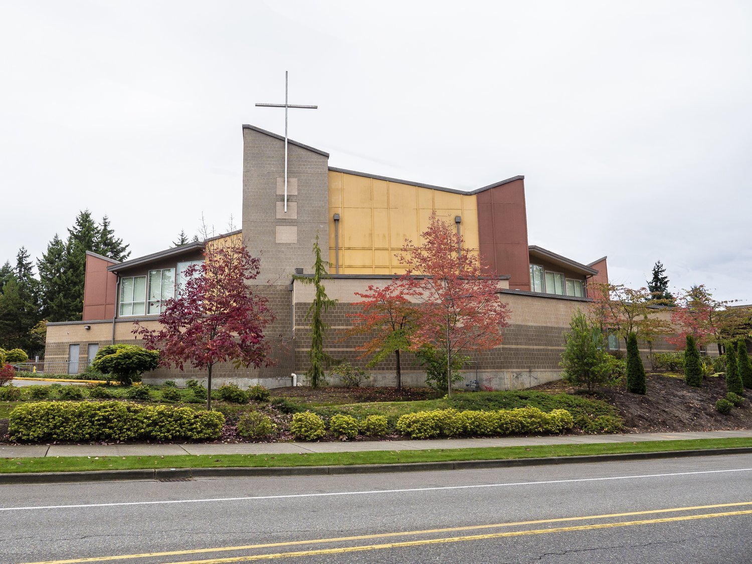 Modern church building with a cross on top, surrounded by landscaped trees and shrubs, located next to a road.