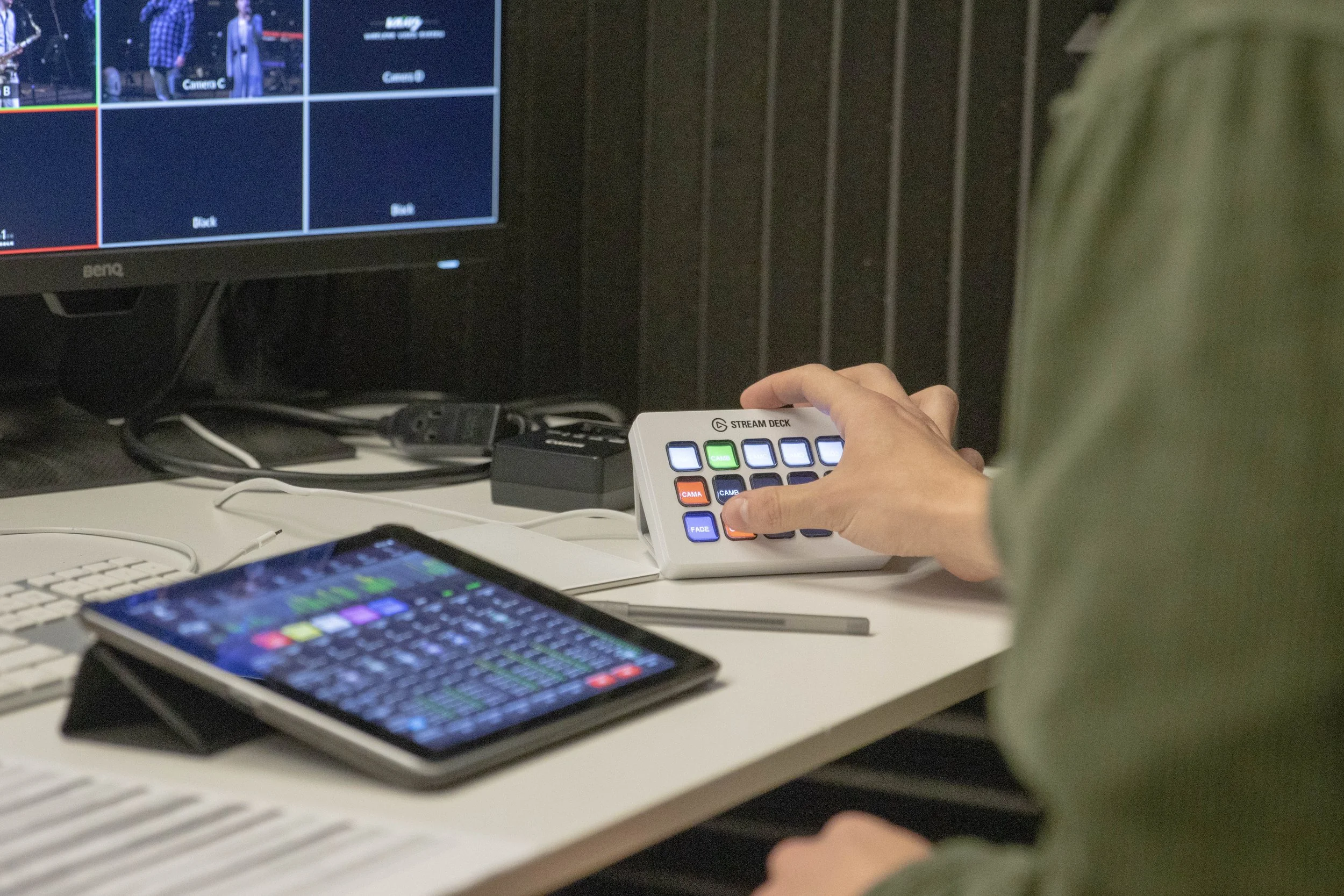 Person operating a stream deck with multiple buttons at a desk, with a monitor displaying video feeds and a tablet showing software controls.
