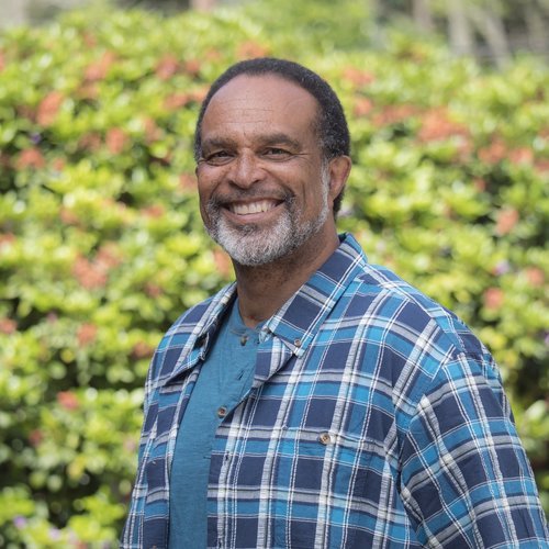 Man smiling outdoors in front of green shrubbery, wearing a blue plaid shirt and turquoise t-shirt.