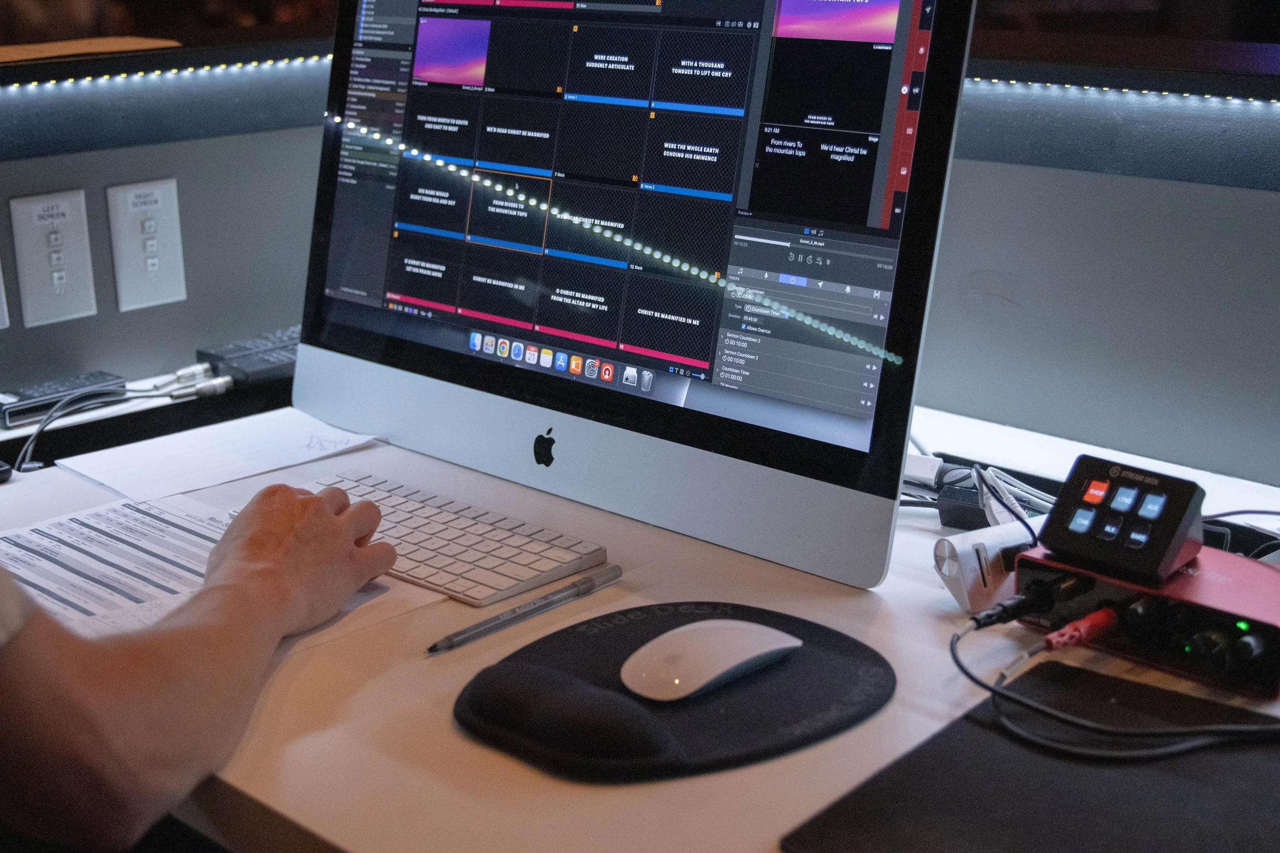 Person using an Apple iMac with a digital software interface displayed on the screen, surrounded by a keyboard, mouse, paper documents, and audio equipment on a desk.