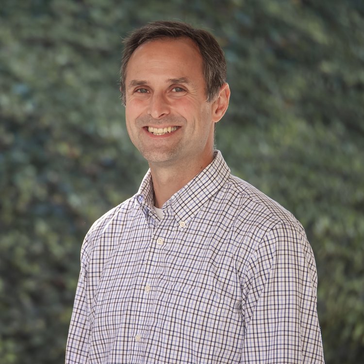 Smiling man in a checked shirt standing outdoors with a blurred green background.