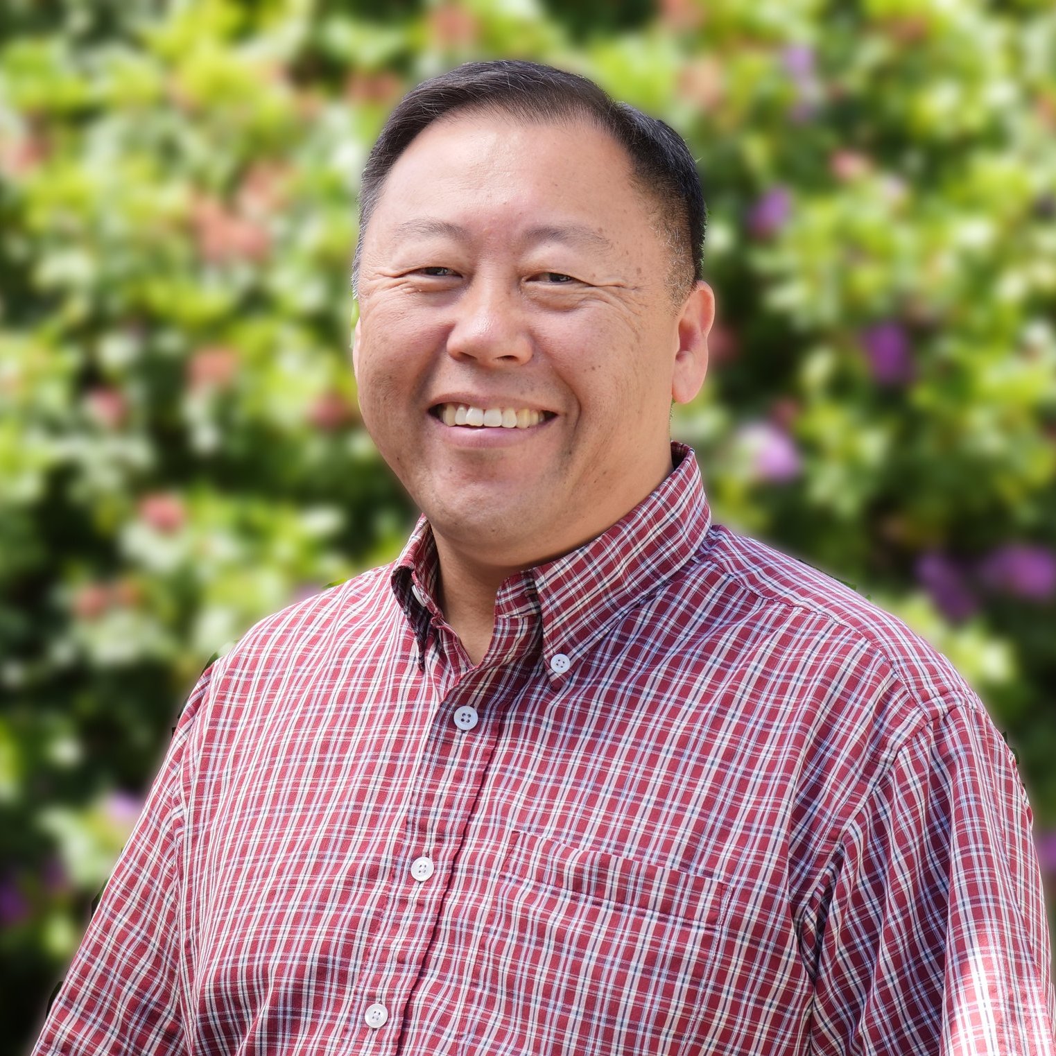 Smiling person in a red plaid shirt in front of a blurred background of green foliage and flowers.