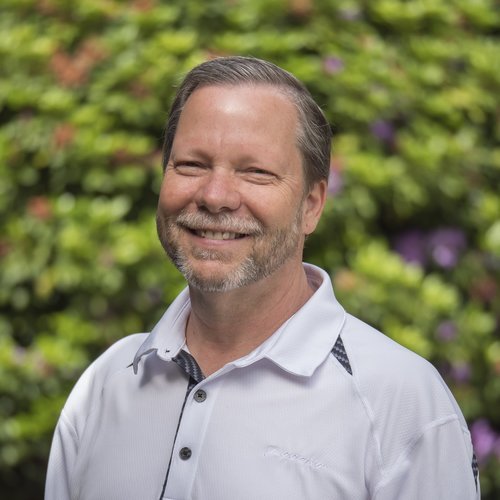 Man in a white shirt smiling against a blurred nature background with greenery and purple flowers.