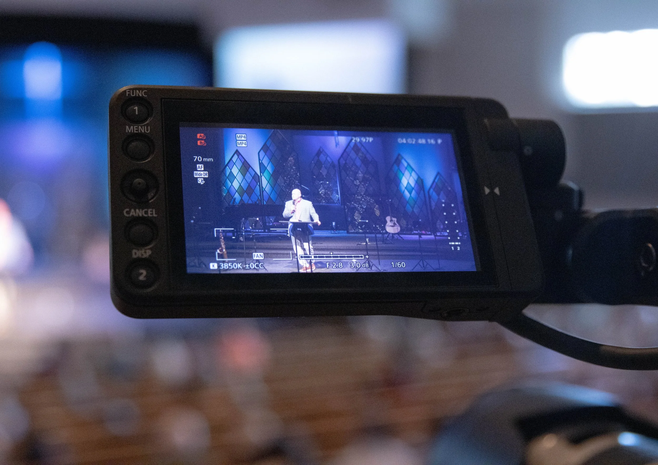 Camera viewfinder showing a person speaking at a podium on stage with musical instruments and modern backdrop.