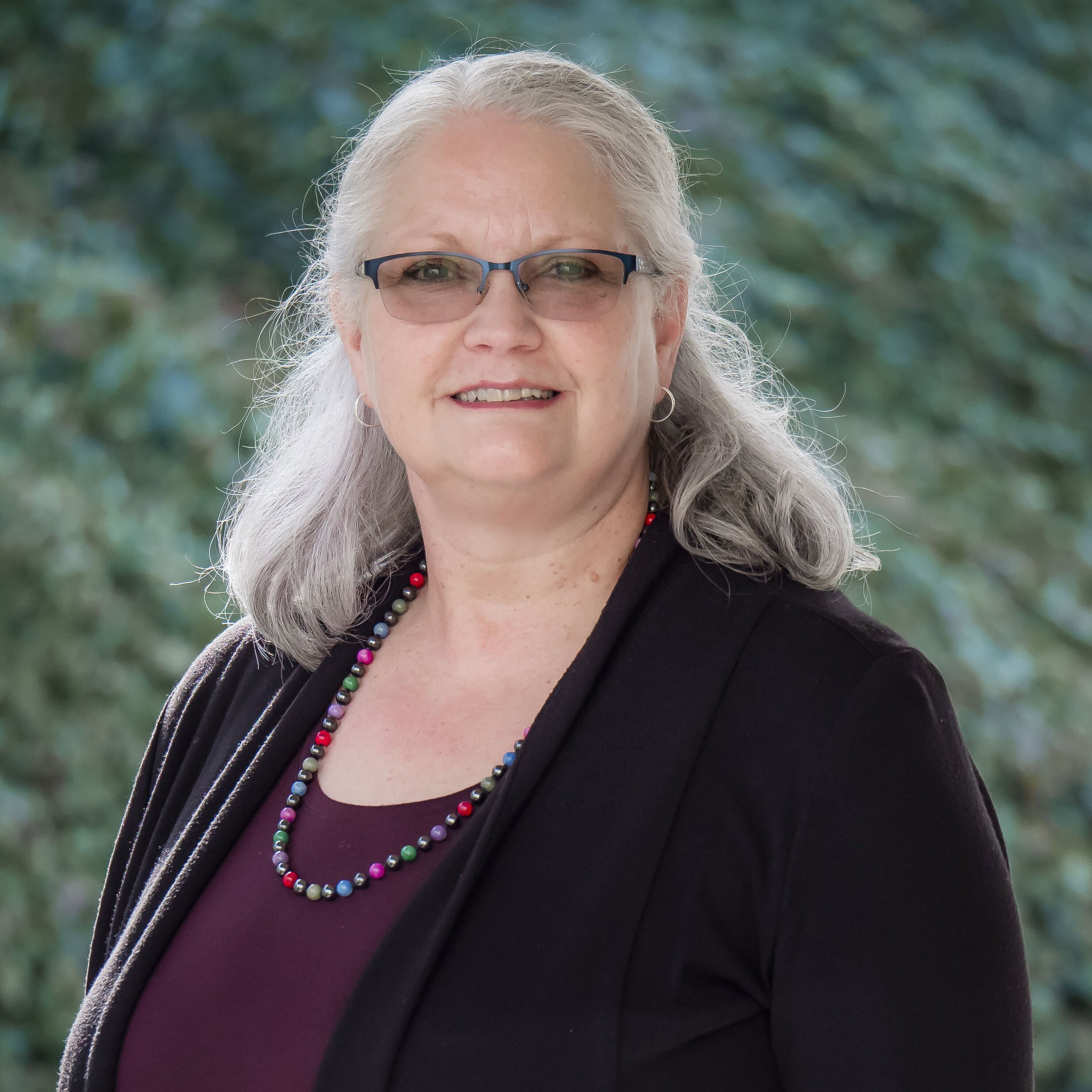 Woman with glasses and long gray hair wearing a necklace and black jacket, standing outdoors against a blurred green background.