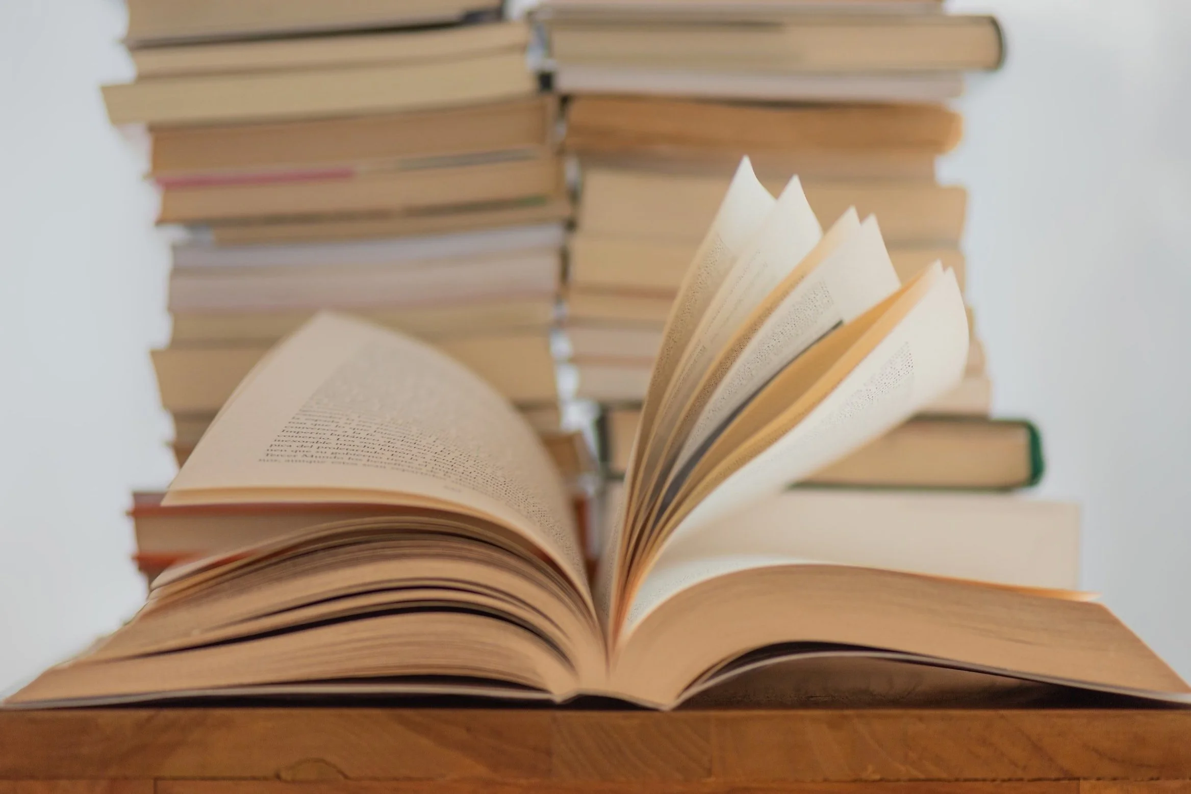 Open book on a wooden surface with a background of many stacked books.