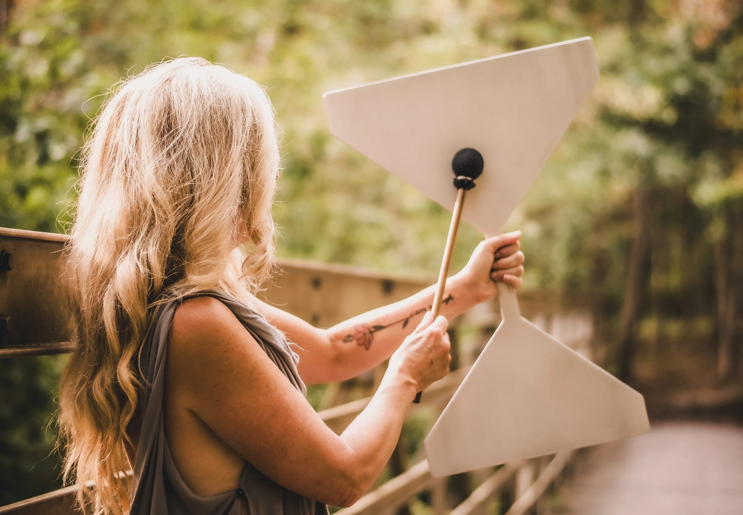 A woman with long blonde hair holding a white sign while standing outdoors near a wooden fence, with a background of blurred green trees.