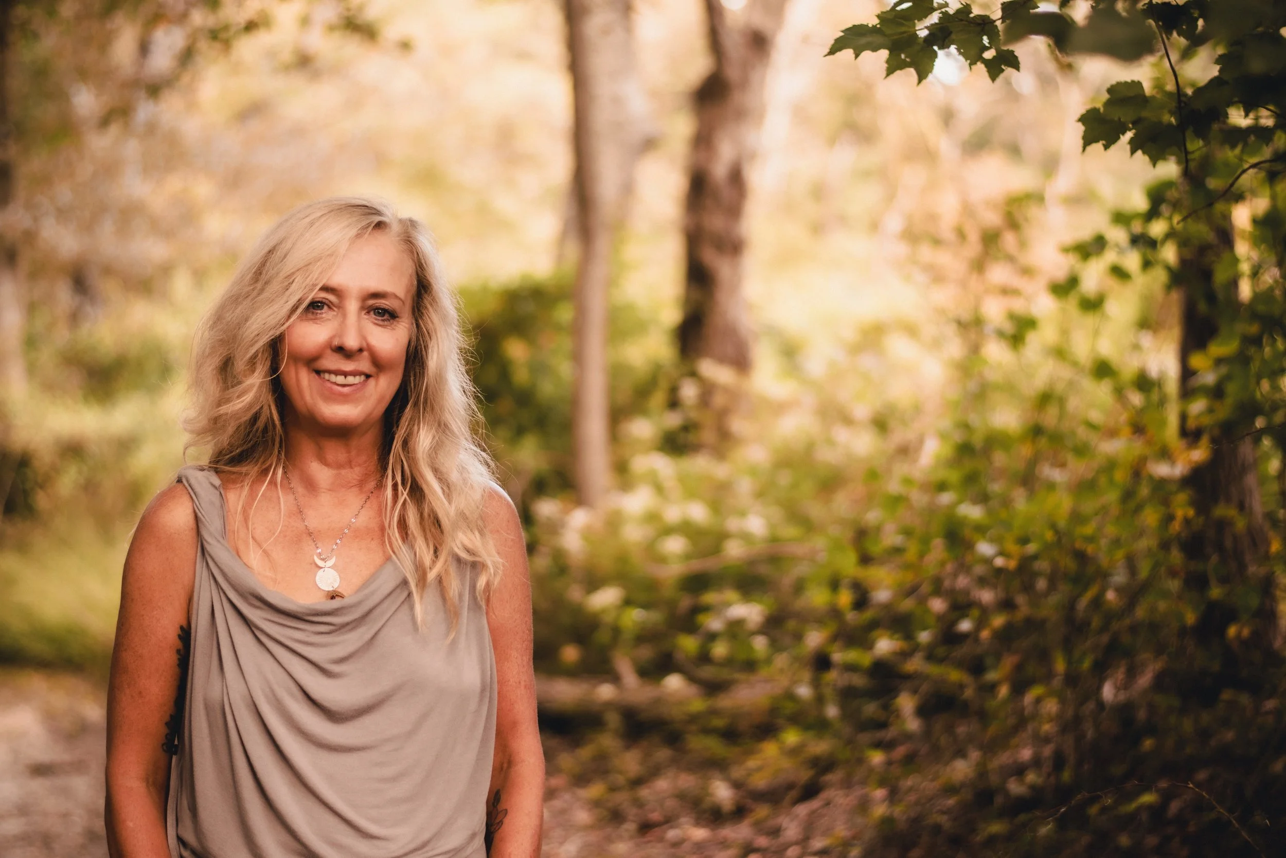 A woman with long blonde hair smiling outdoors in a wooded area with green and yellow foliage.