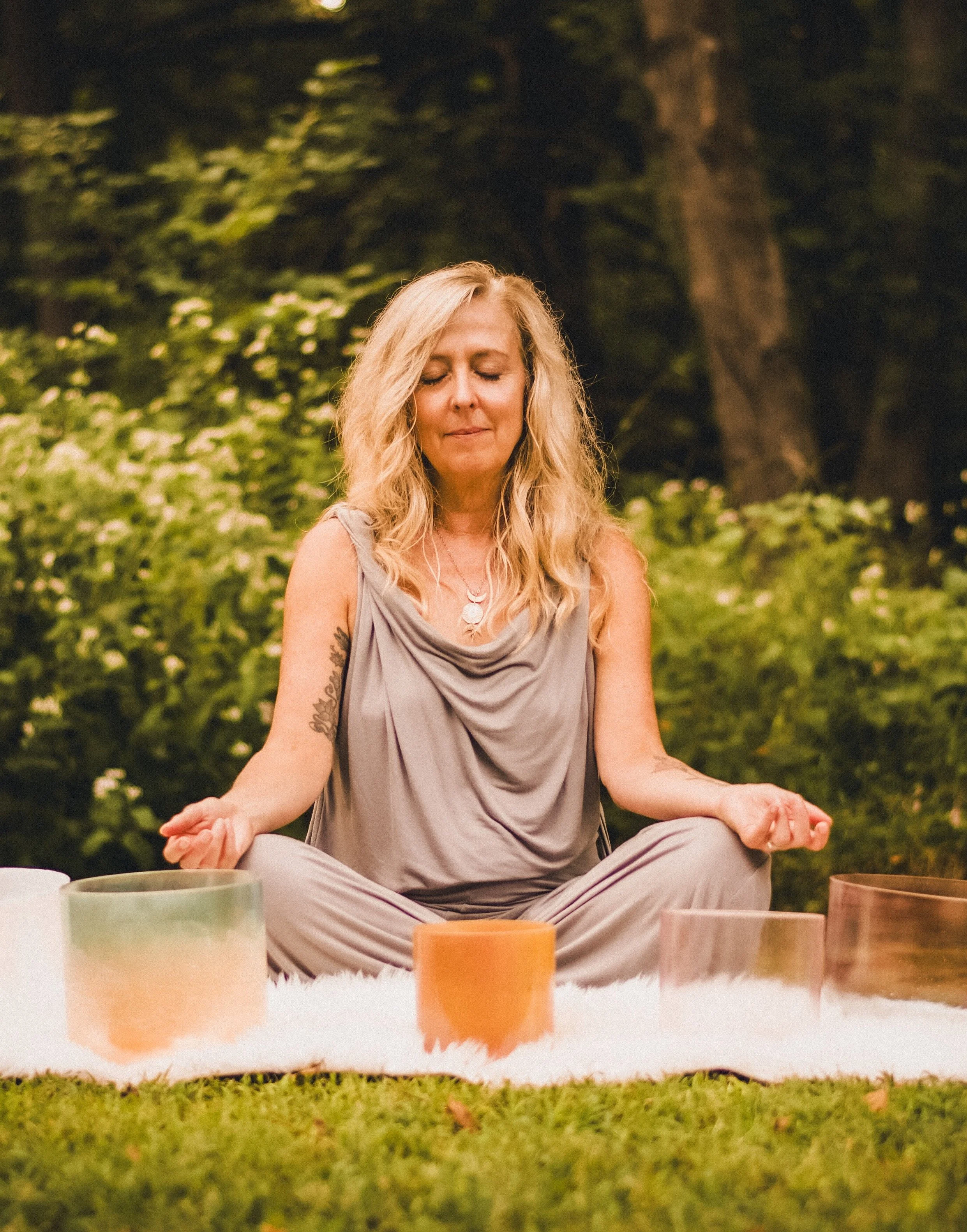 A woman with long blonde hair sitting cross-legged outdoors on grass, meditating with eyes closed, surrounded by various colorful cups and bowls, in a lush green setting during daytime.