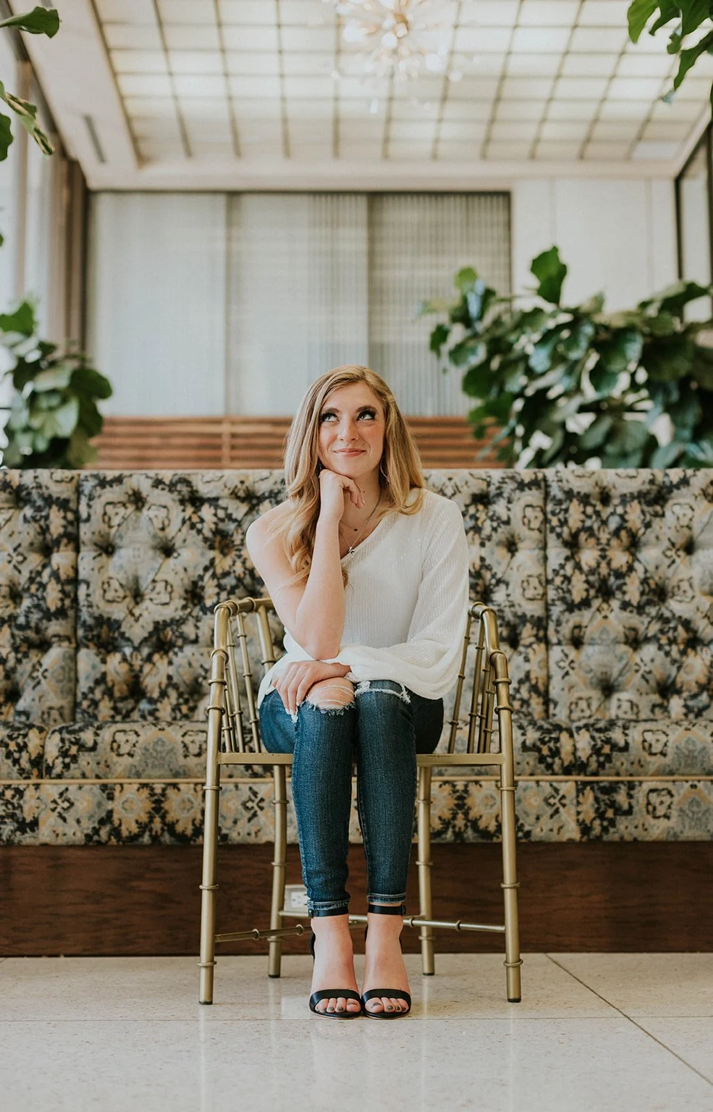 A young woman with long blonde hair sitting on a brass chair in a stylish indoor setting with a floral patterned sofa and green plants behind her. She is wearing a white sweater, ripped jeans, and black high-heeled sandals, and she is smiling while r