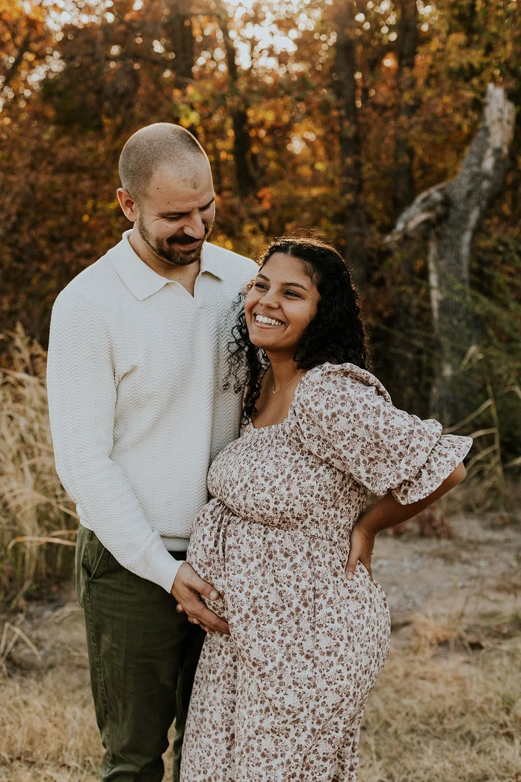 A happy couple, a pregnant woman and a man, standing outdoors in a natural setting during fall, with trees and foliage in the background.