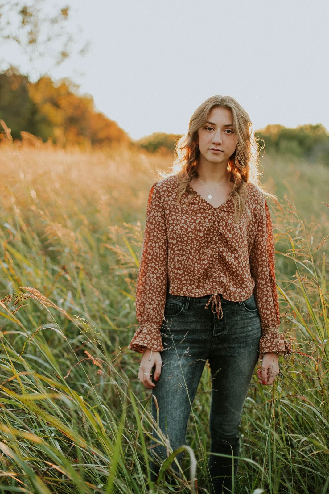 A young woman with wavy blonde hair standing in a grassy field during sunset, wearing a brown patterned blouse and dark jeans.