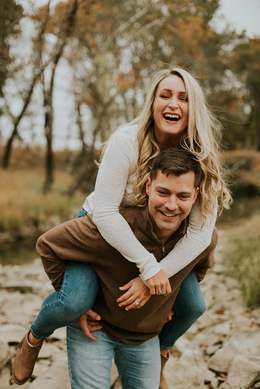 A smiling woman with long blonde hair riding piggyback on a man with short dark hair outdoors in a wooded area during autumn.