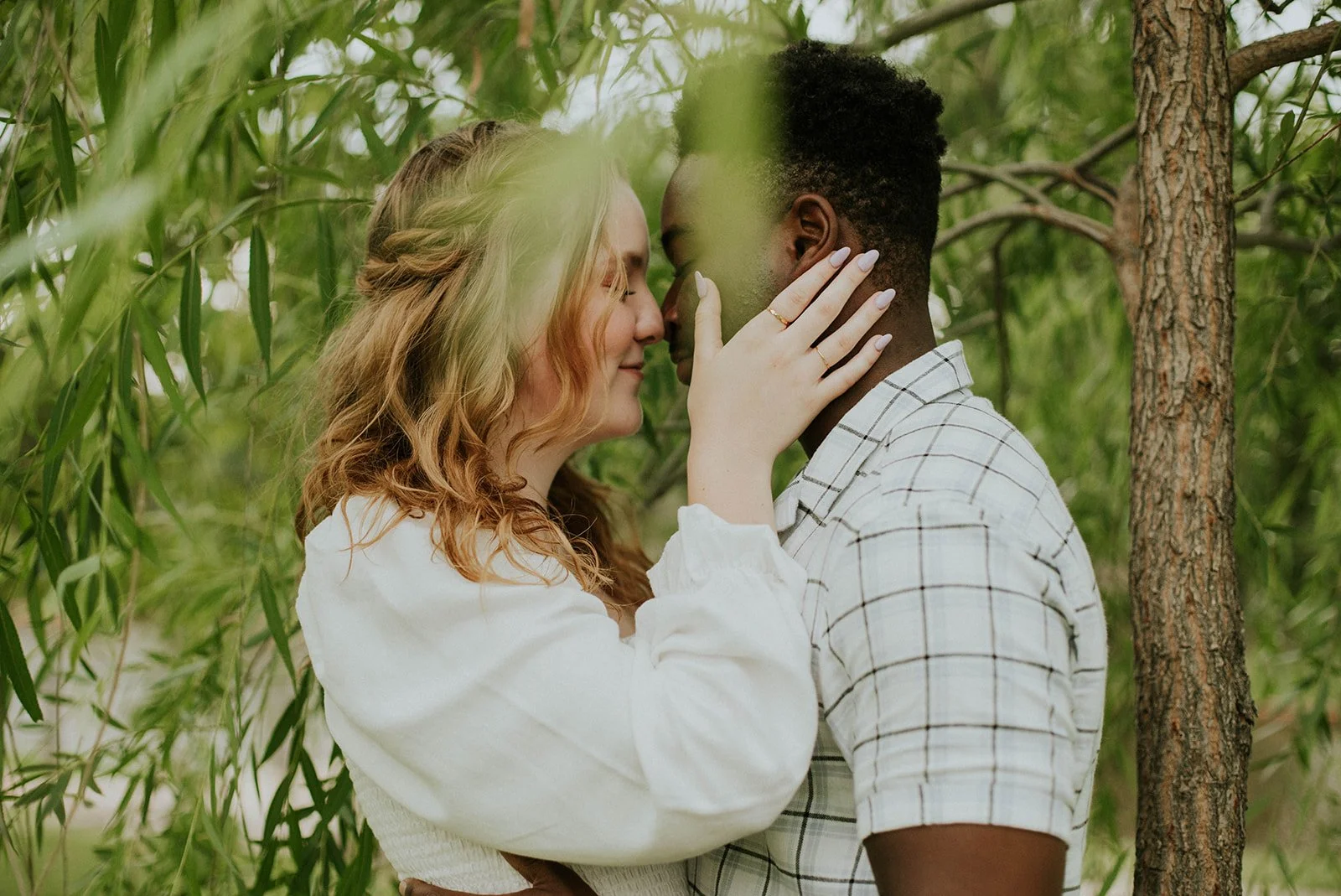 A couple standing close together outdoors, touching foreheads and smiling, surrounded by green leaves and trees.