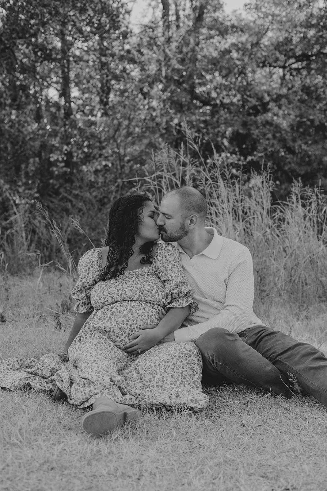A black and white photo of a pregnant woman and a man sitting on the grass, kissing outdoors with trees in the background.